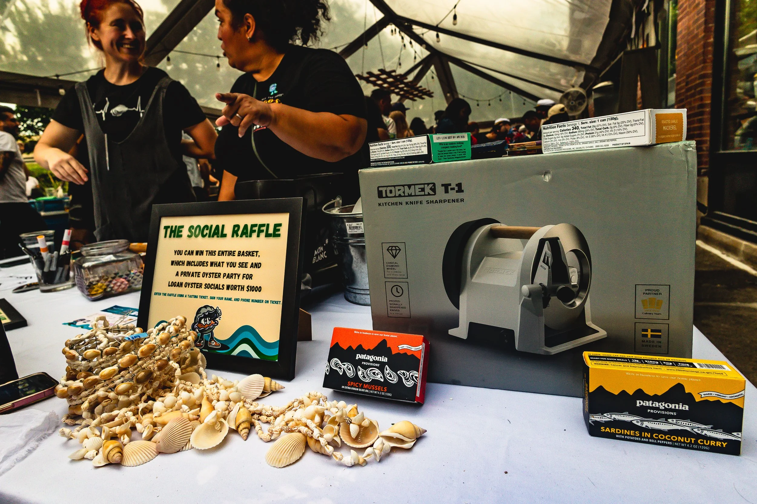 A table at an event with a sign for a social raffle, seashells, a kitchen knife sharpener, canned seafood, and a box of spicy mussels, with two women talking in the background under a tent.