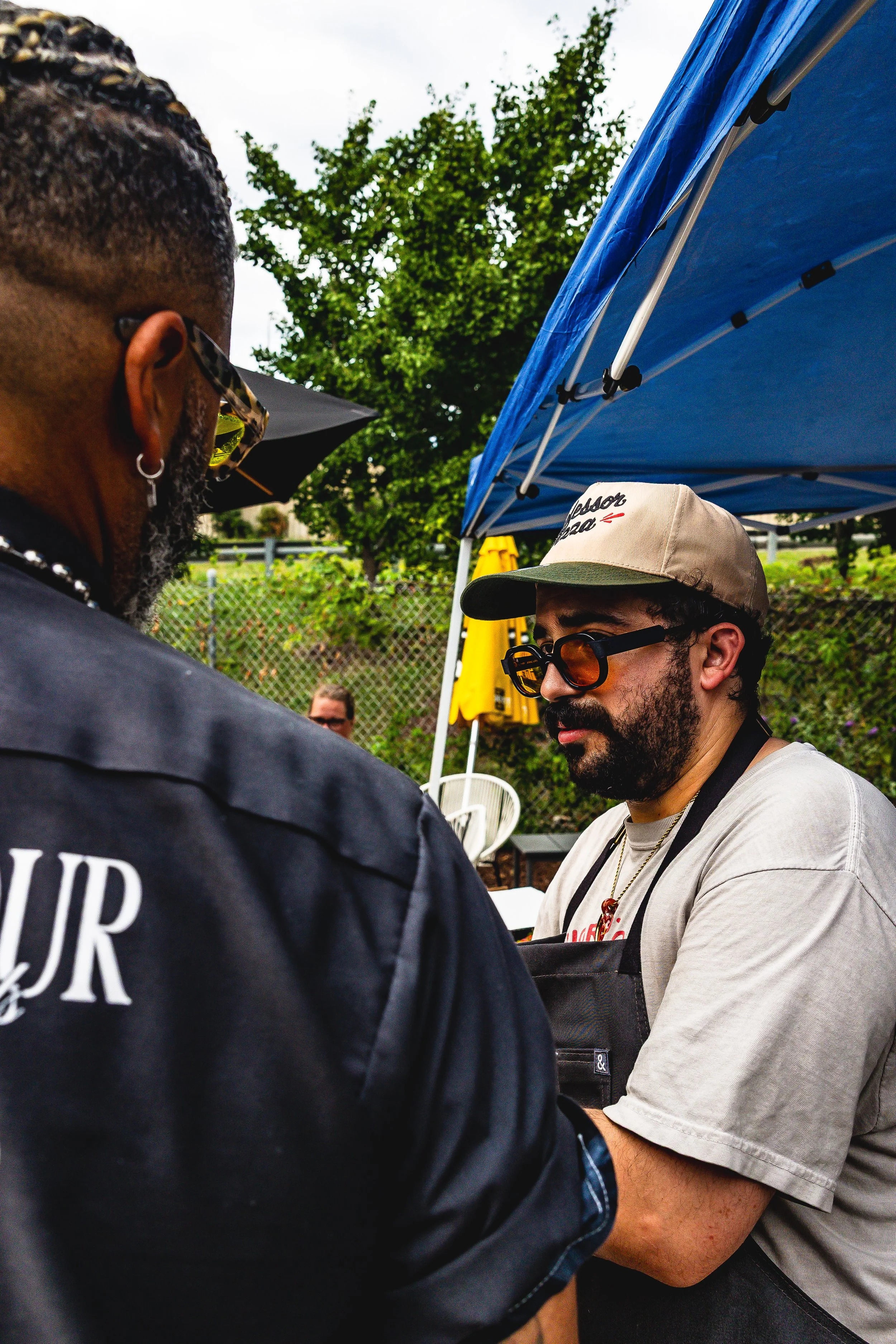 Two men having a conversation outdoors at a gathering, under a blue canopy tent, with trees and a fence in the background.