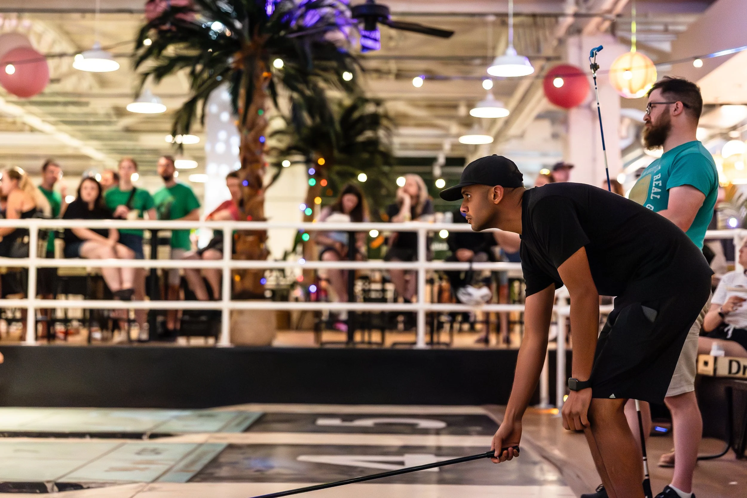 A man with a black cap and black t-shirt crouching down holding a fishing rod-like device, focused on the ground in an indoor setting with a crowd of people seated behind a white barrier, decorated with colorful lights and paper lanterns.