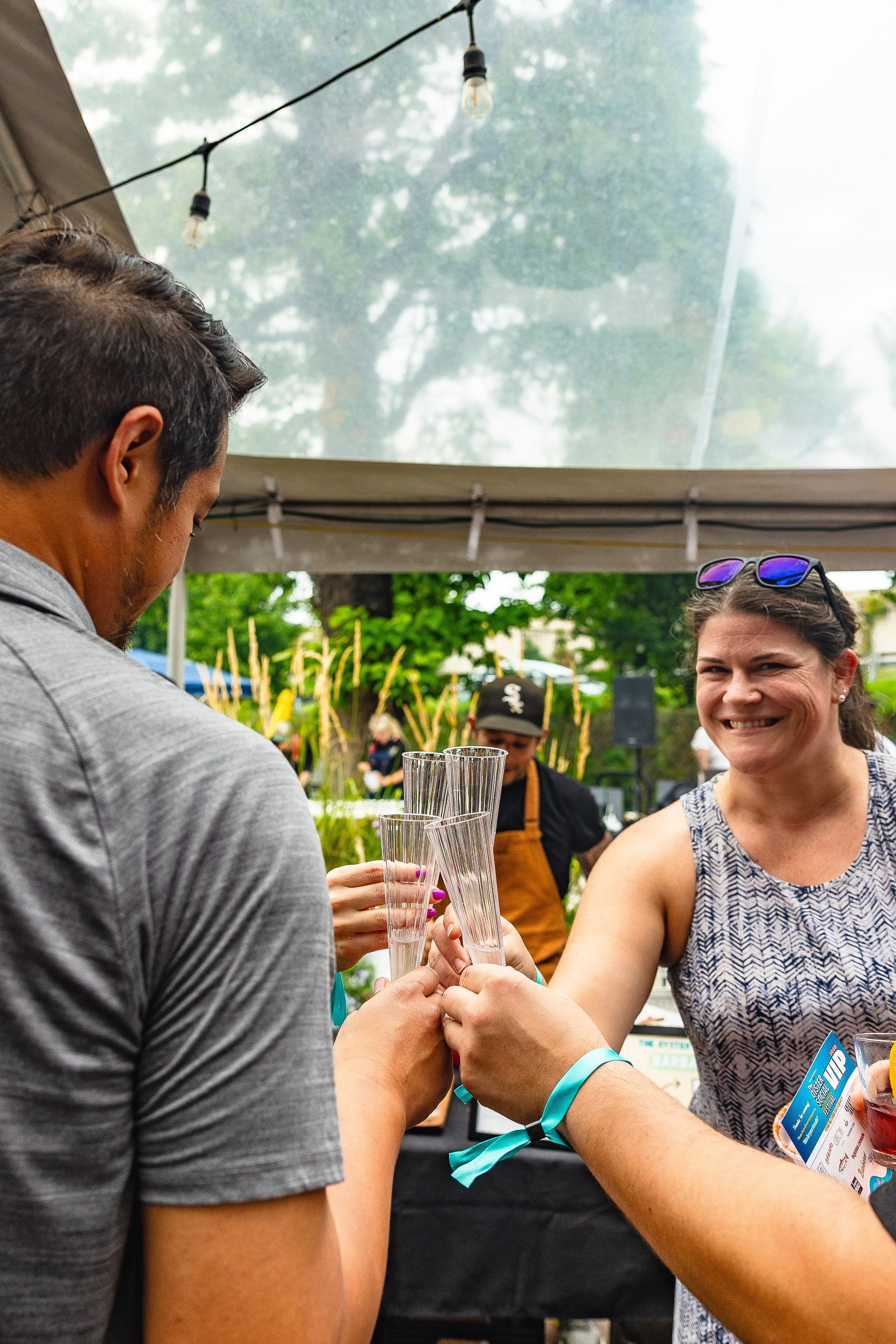People clinking glasses at an outdoor event under a tent with string lights, smiling woman in sleeveless top and sunglasses on her head.