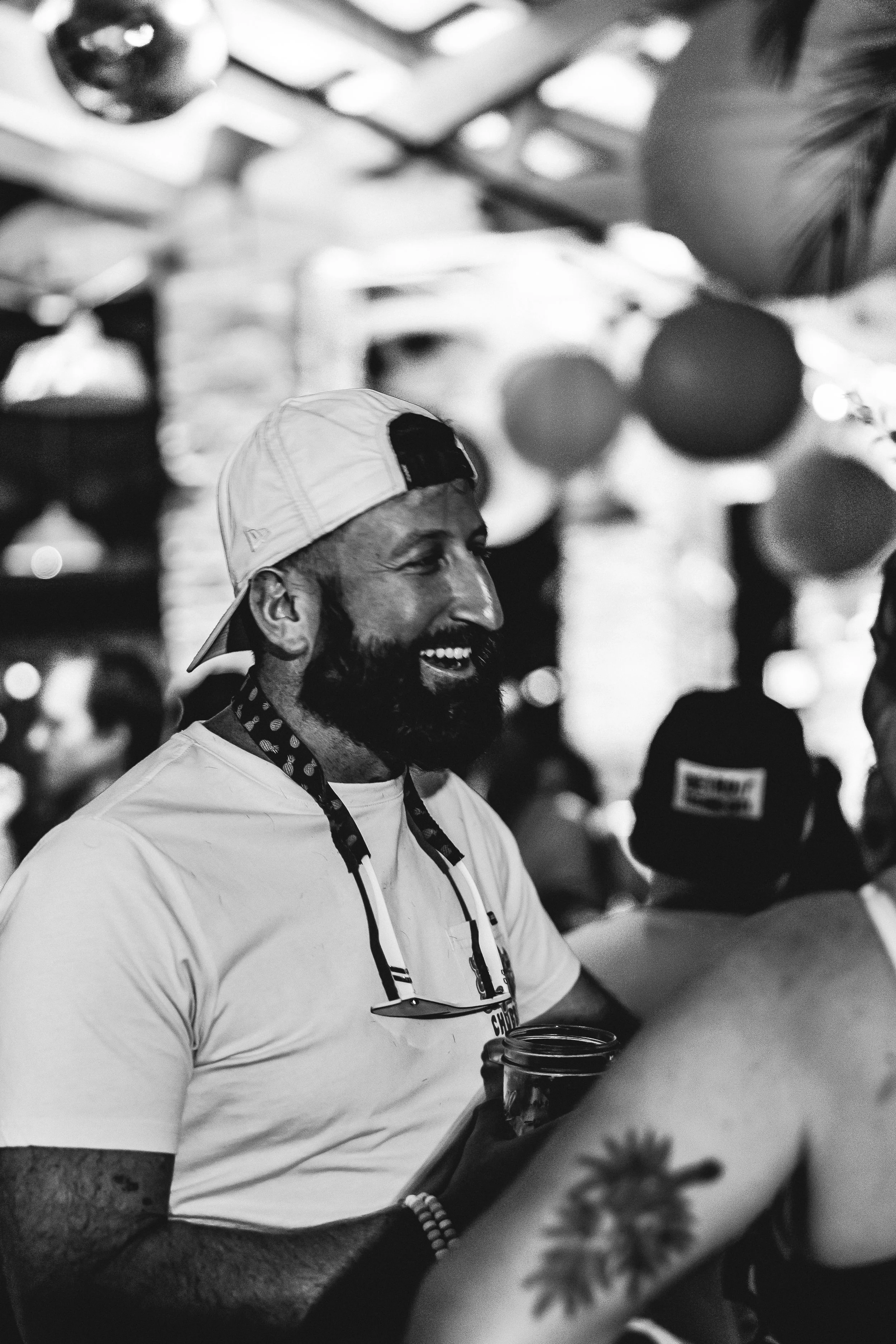 A man with a beard and a baseball cap smiling and holding a drink at a lively event with balloons and people in the background.
