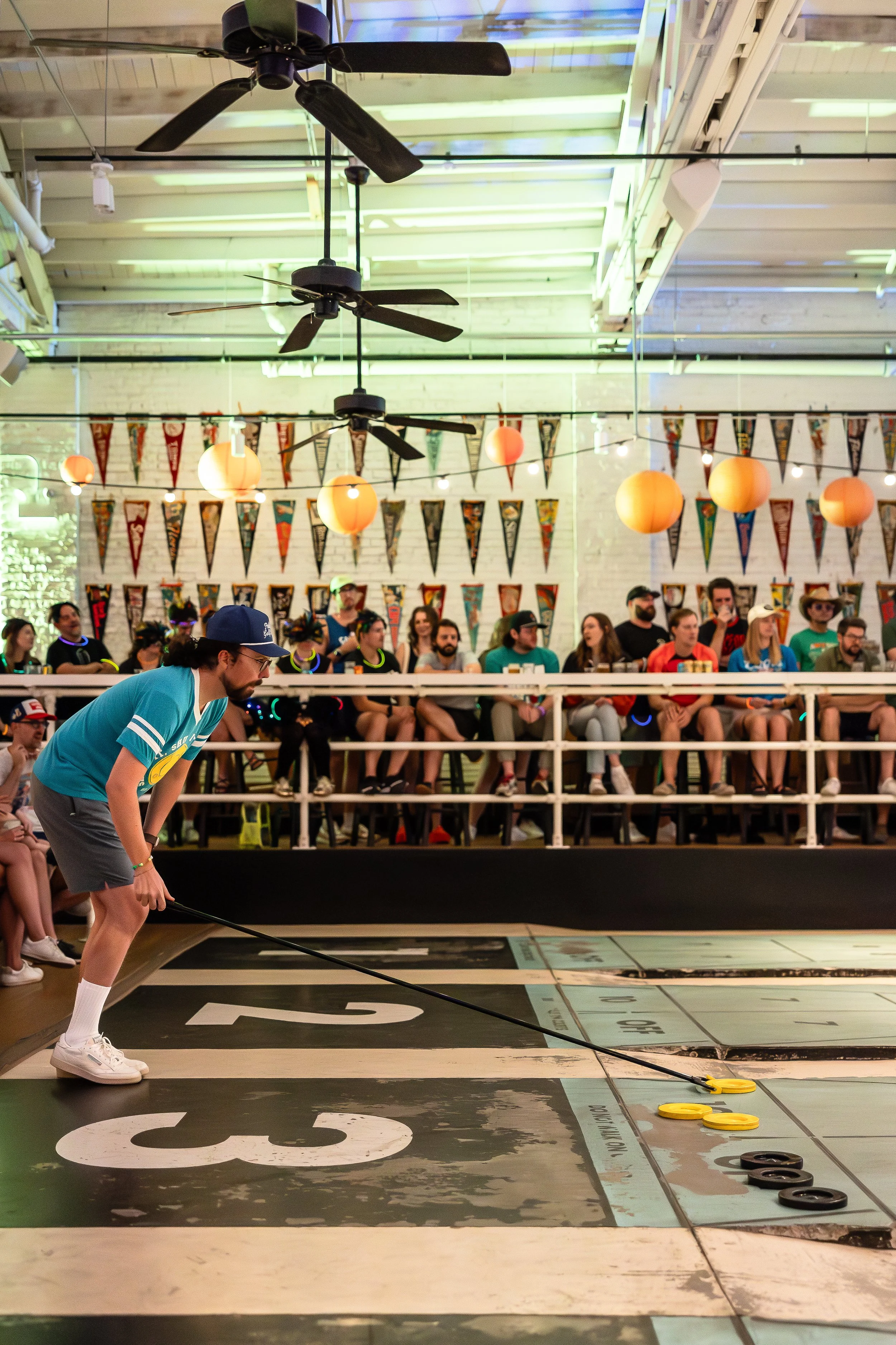 A group of people watching a shuffleboard game in a decorated indoor venue with hanging lanterns and banners.