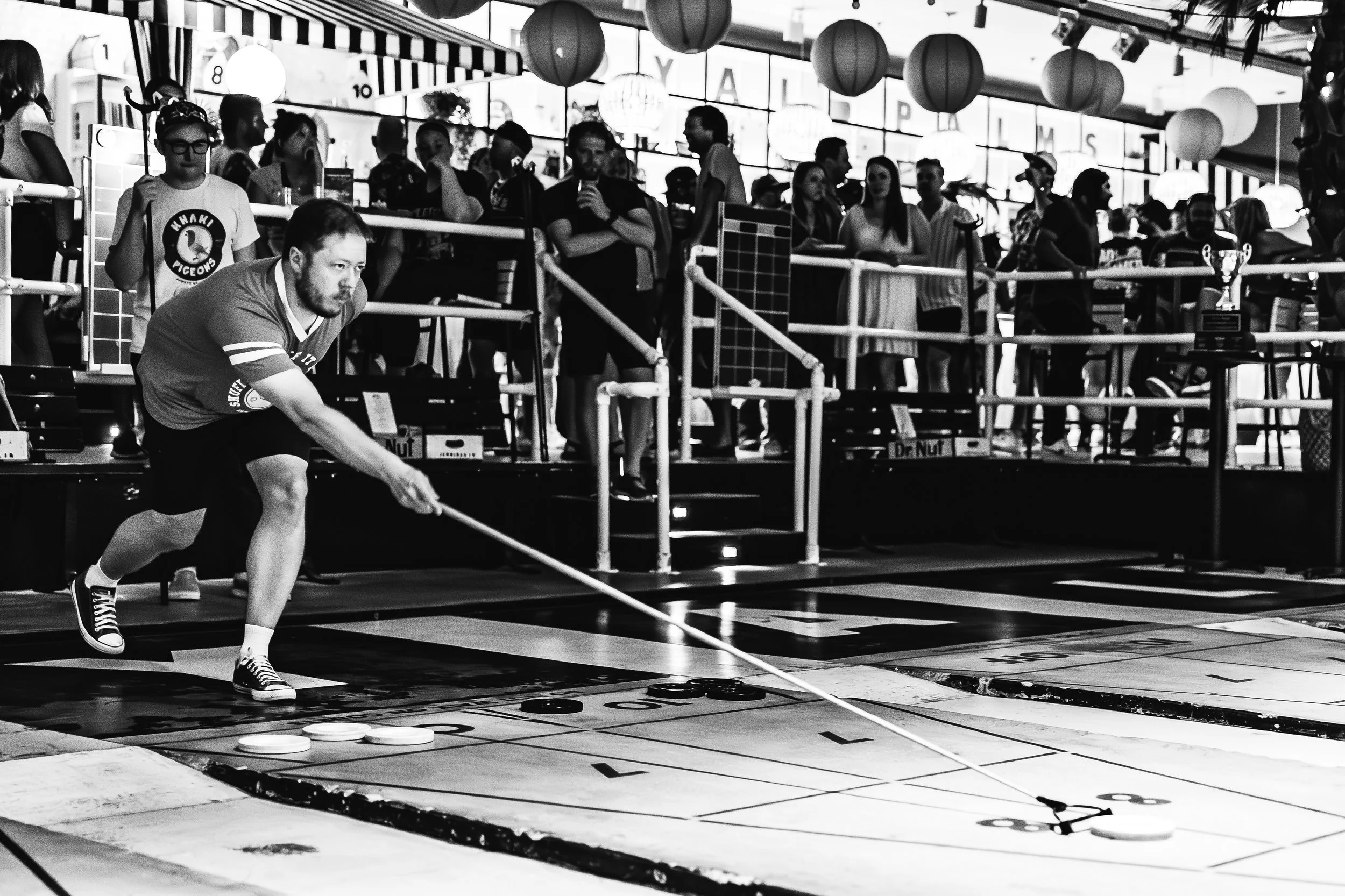 A man playing floor shuffleboard during a competition, with spectators watching in the background at an indoor venue decorated with hanging paper lanterns.