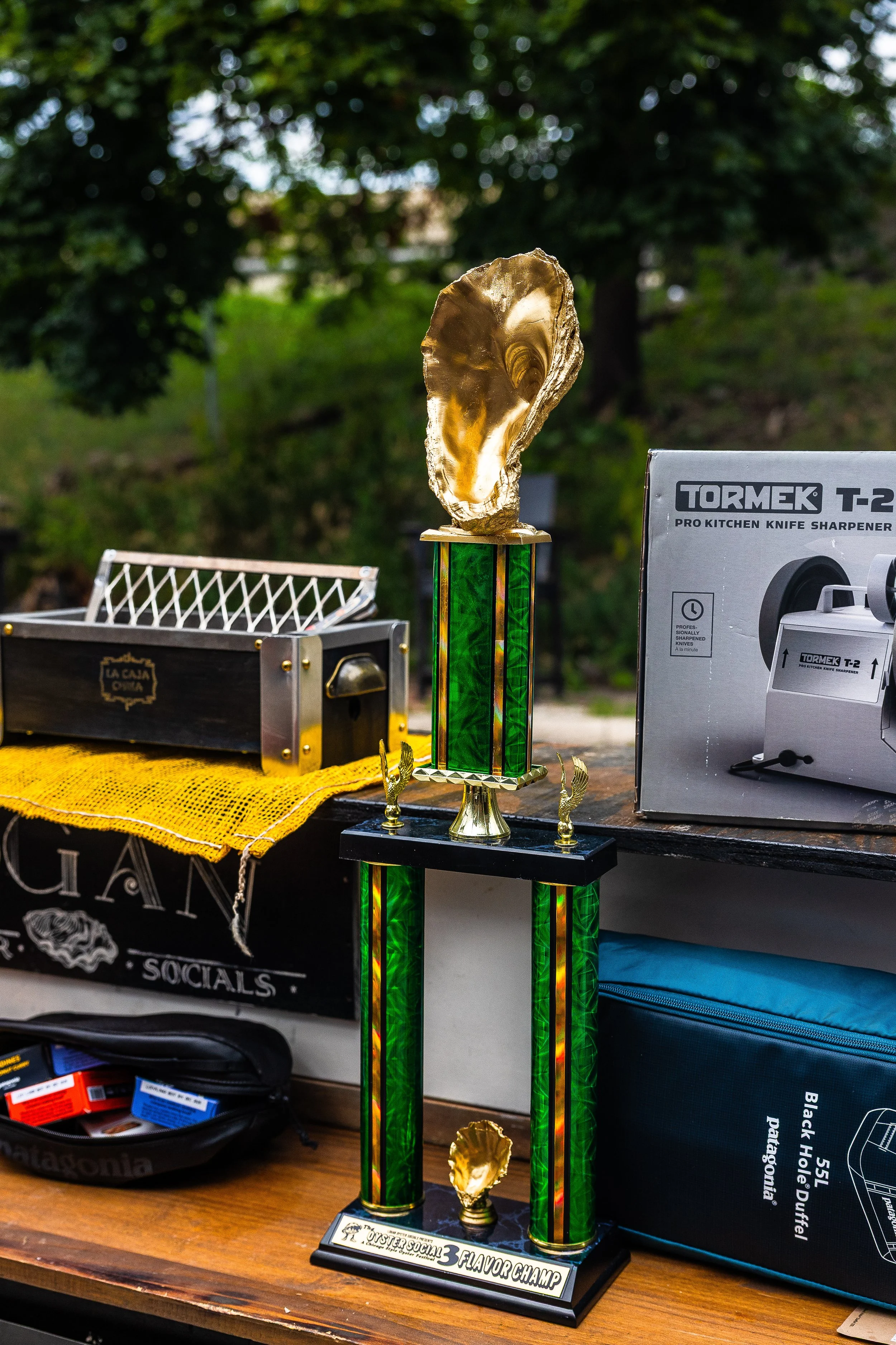 A trophy with a gold oyster shell on top, green and gold columns, on a black base with a plaque, placed on a wooden surface.
