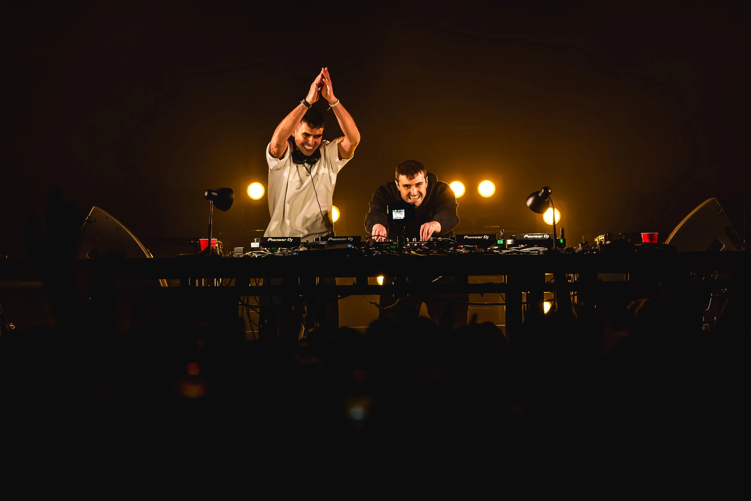Fred Again and Sammy Vigil performing on stage with DJ equipment, illuminated by yellow stage lights in the background, one is clapping and the other is smiling while adjusting controls.