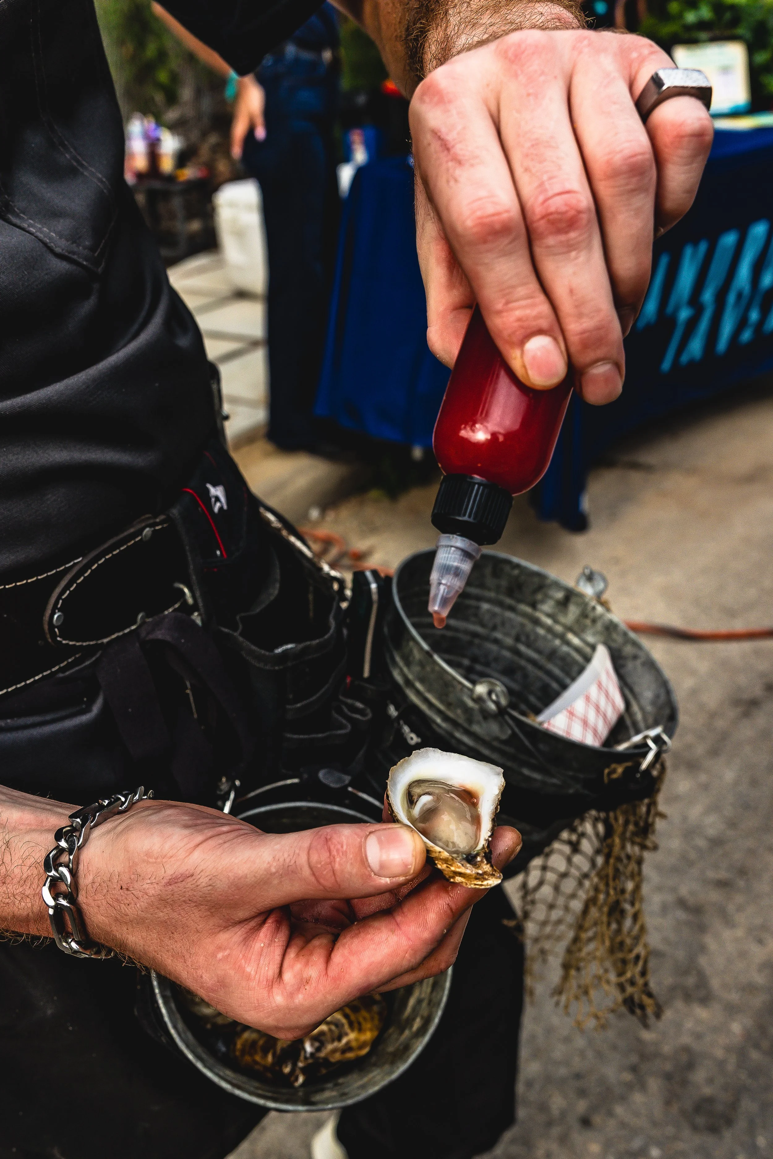 A person holding a half-open oyster with an oyster knife. The person's other hand is squeezing a red sauce bottle over the oyster. The person is wearing a silver bracelet and a black wristwatch.