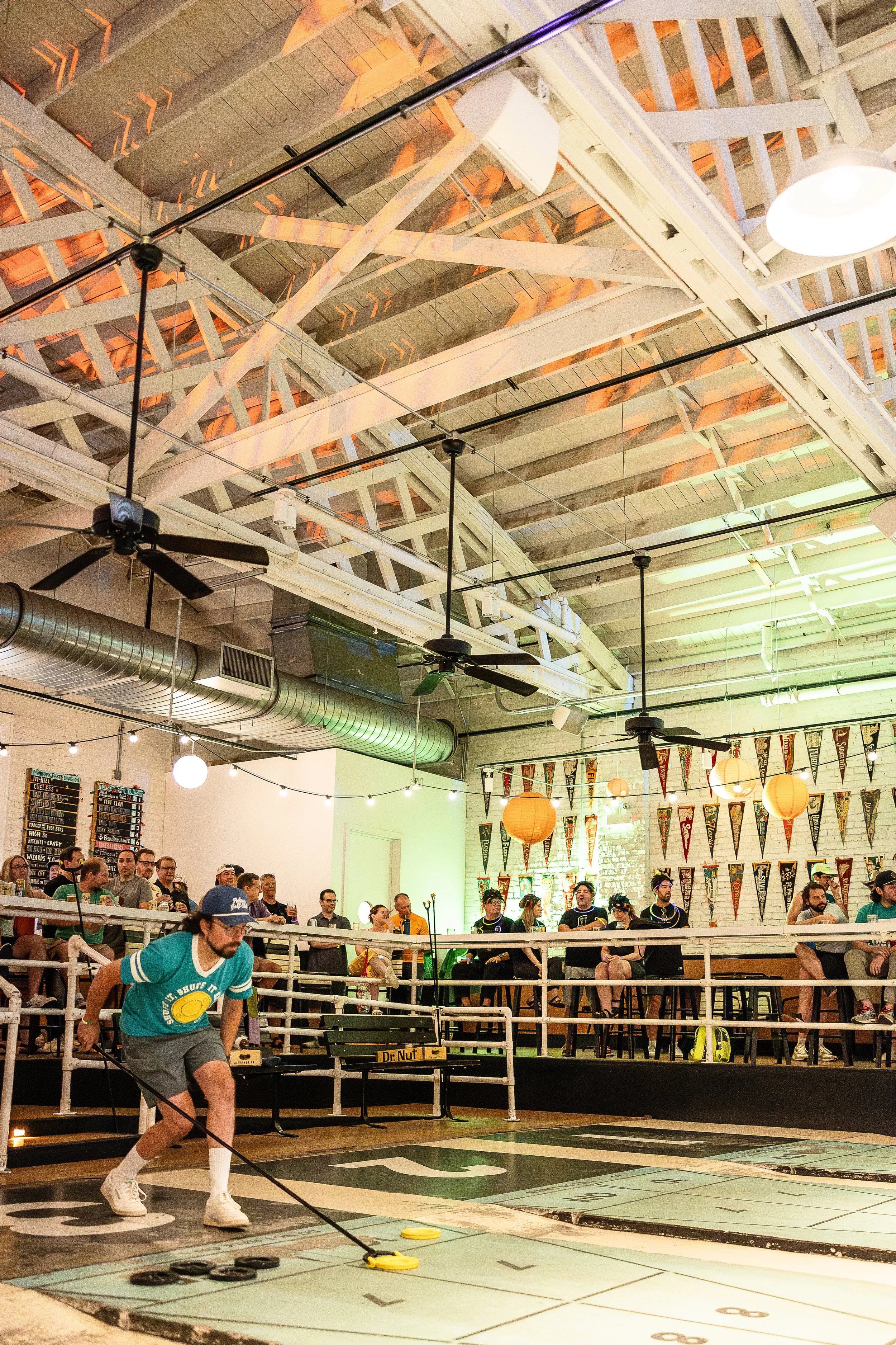 People playing shuffleboard inside a decorated indoor space with hanging paper lanterns and banners, some spectators watching, and a man in foreground preparing to slide a yellow disc.