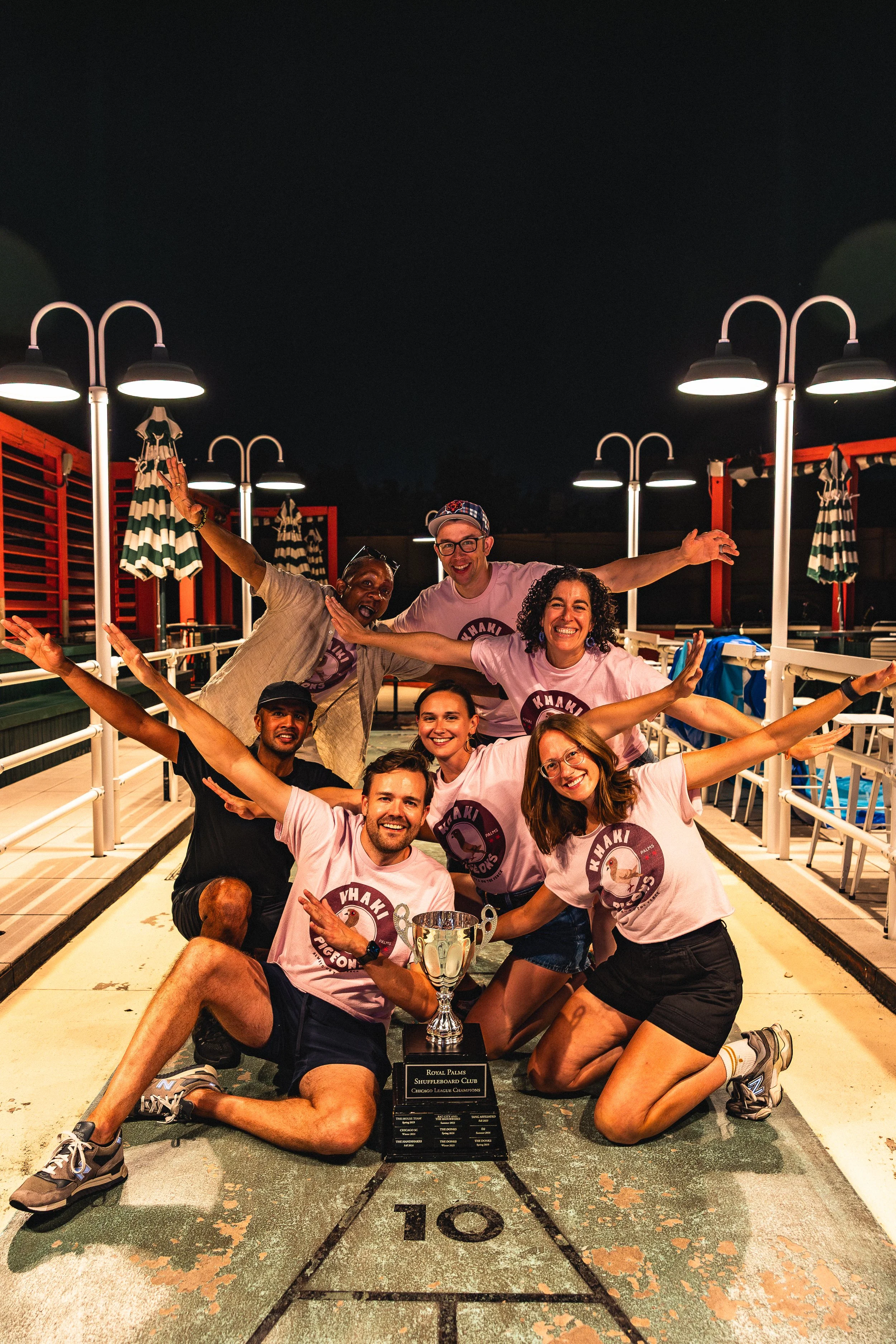 Group of six people celebrating on a shuffleboard court at night, holding a trophy and smiling, with some members wearing matching pink t-shirts that say 'Wiaki Pigeons.'