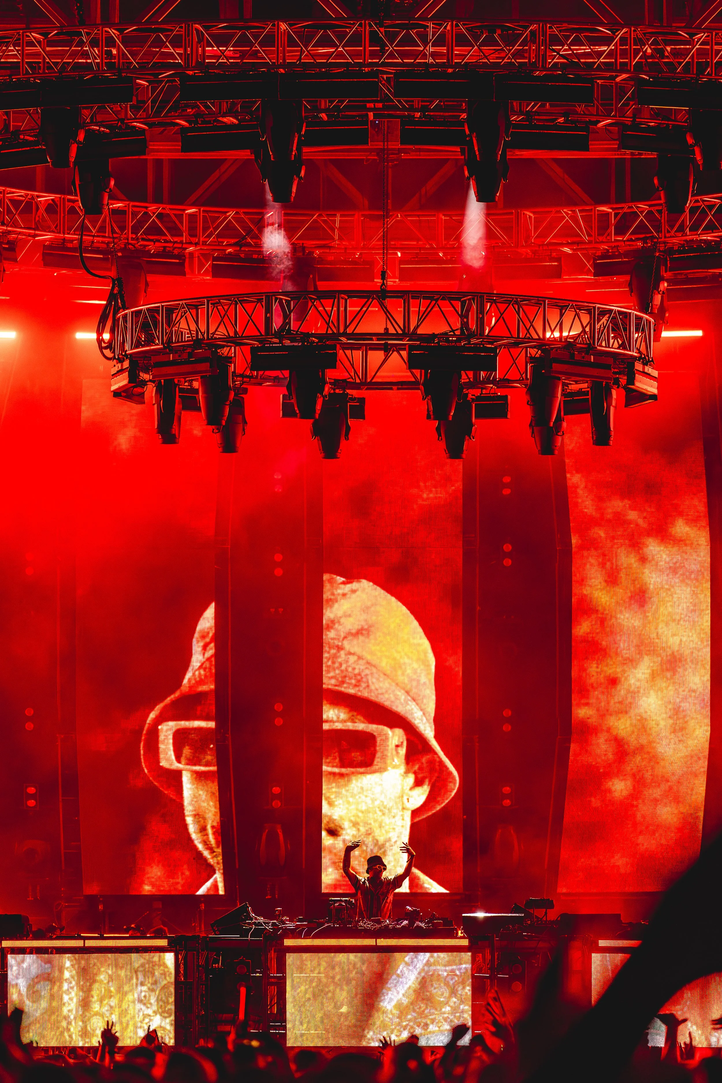 A DJ performs on stage at a concert with red lighting and a large digital screen displaying a stylized person's face wearing glasses and a bucket hat.