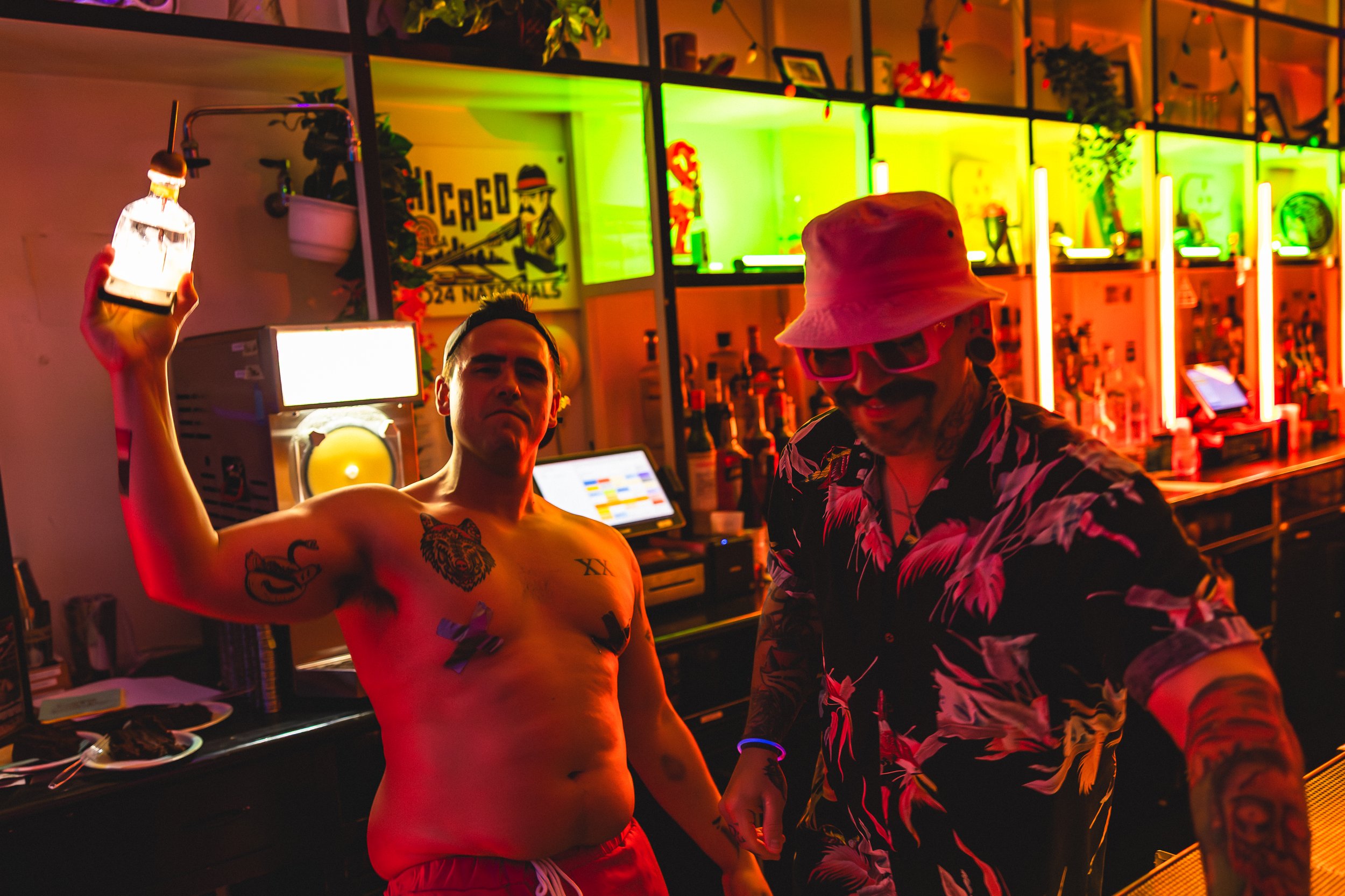 Two men in a colorful bar, one shirtless with tattoos and holding a lantern, the other wearing a floral shirt, hat, and sunglasses, with bottles and neon lights in the background.