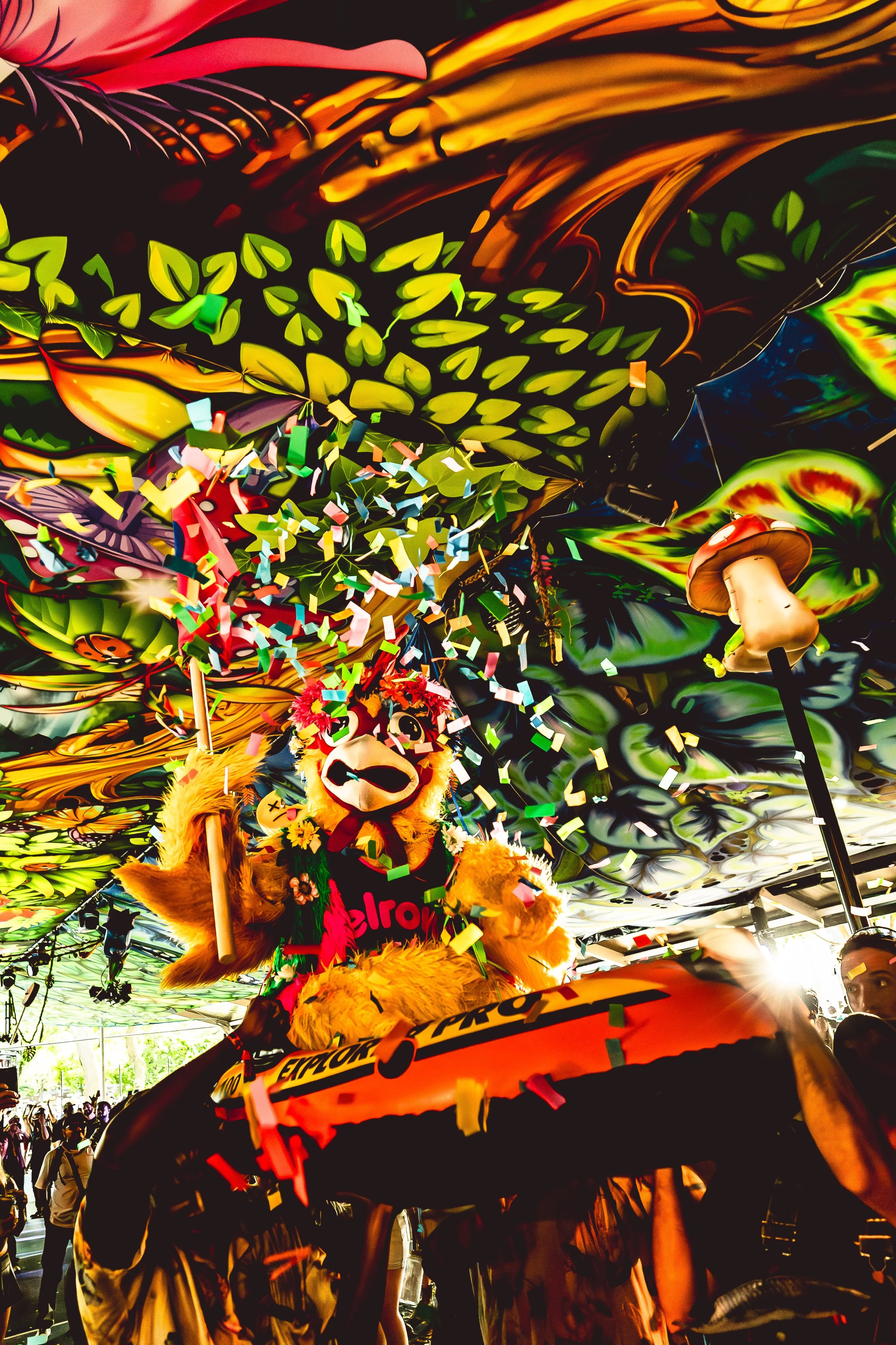 A colorful parade scene with a mascot in a lion costume wearing a flower crown and rainbow scarf, on a float decorated with orange and black, surrounded by confetti and a vibrant, abstract, psychedelic ceiling background.