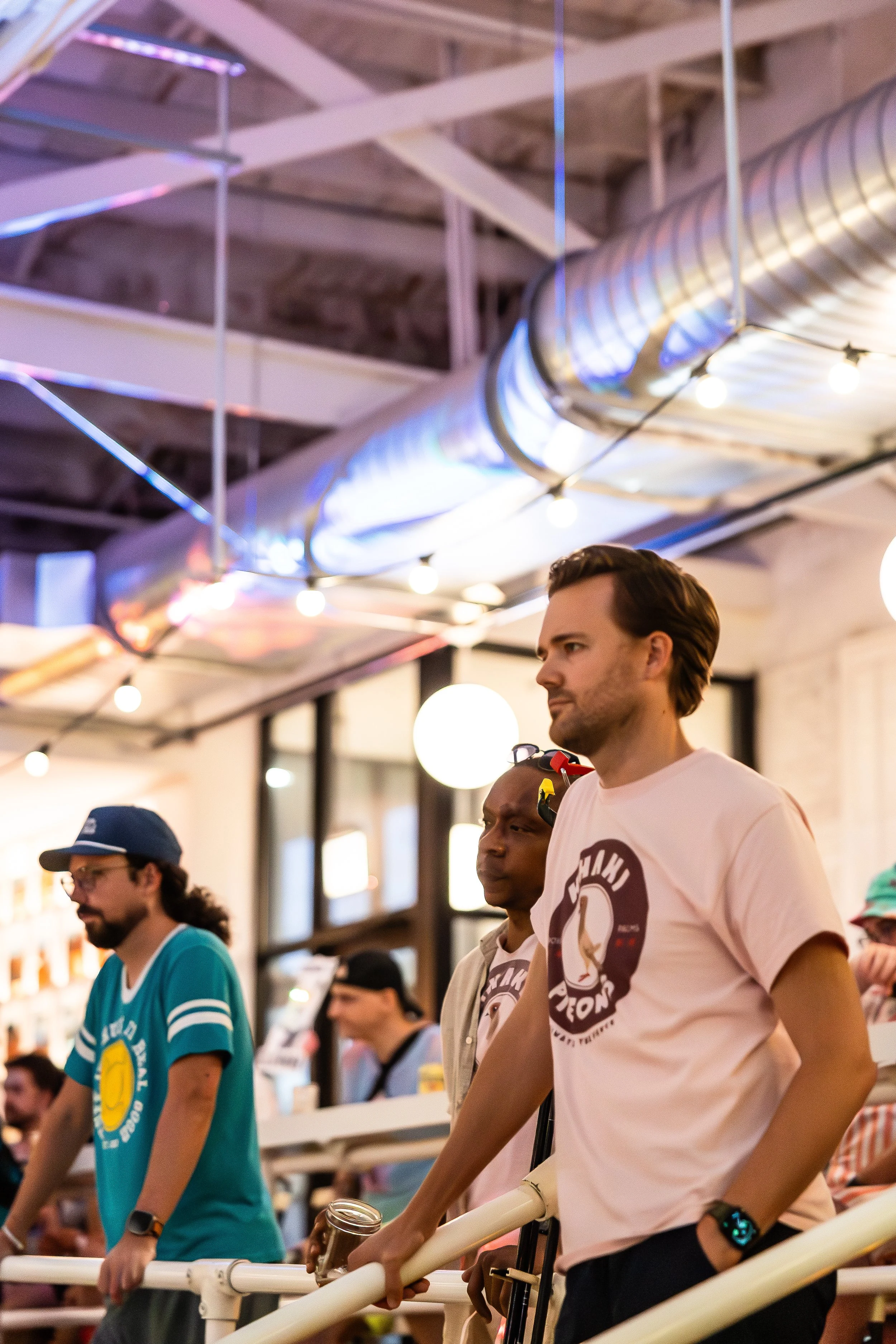 People standing indoors, wearing casual clothes, under exposed ceiling pipes and string lights, possibly at a social or recreational event.