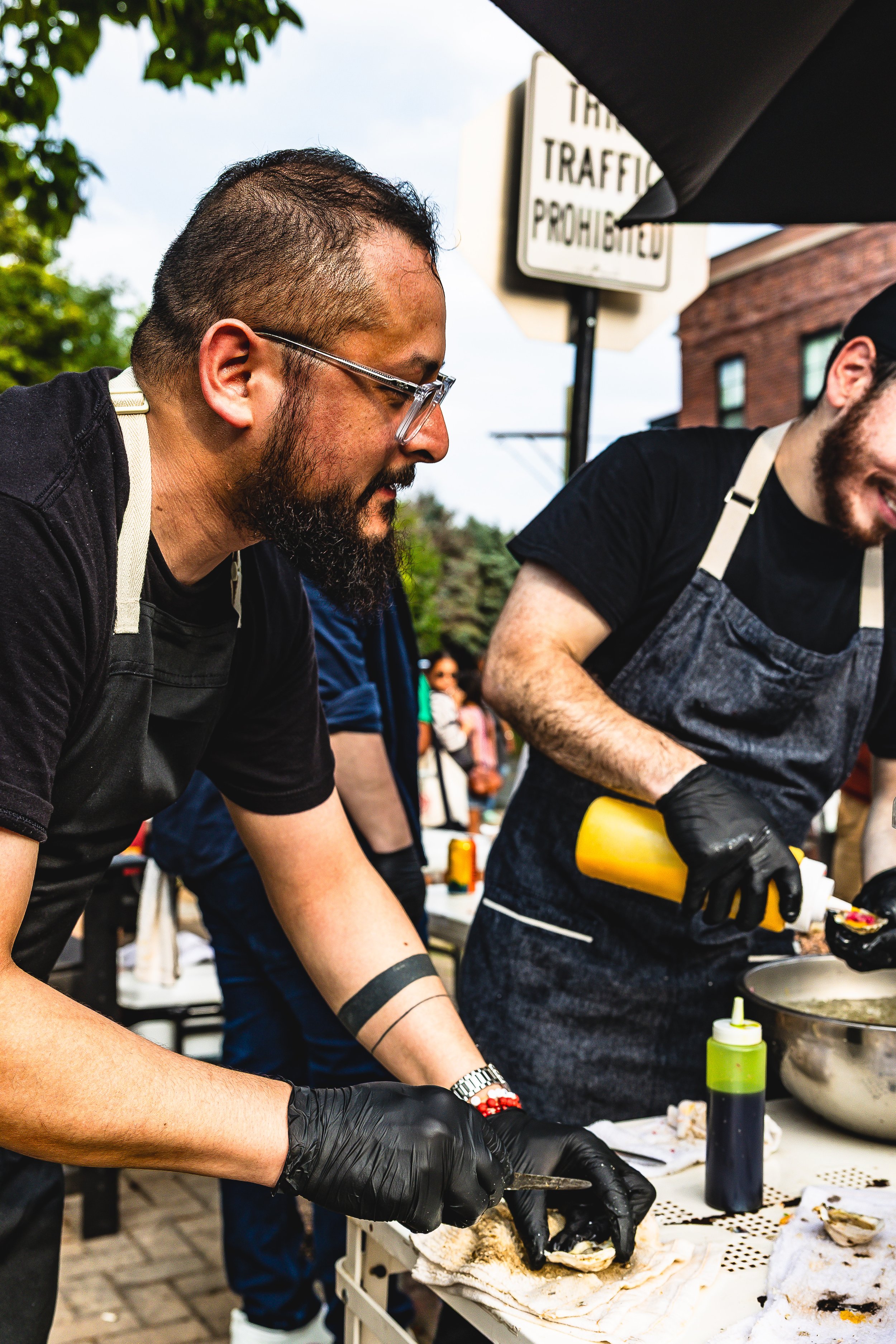 Two men wearing black shirts, aprons, and black gloves preparing food at an outdoor event, with people in the background and a sign about traffic.
