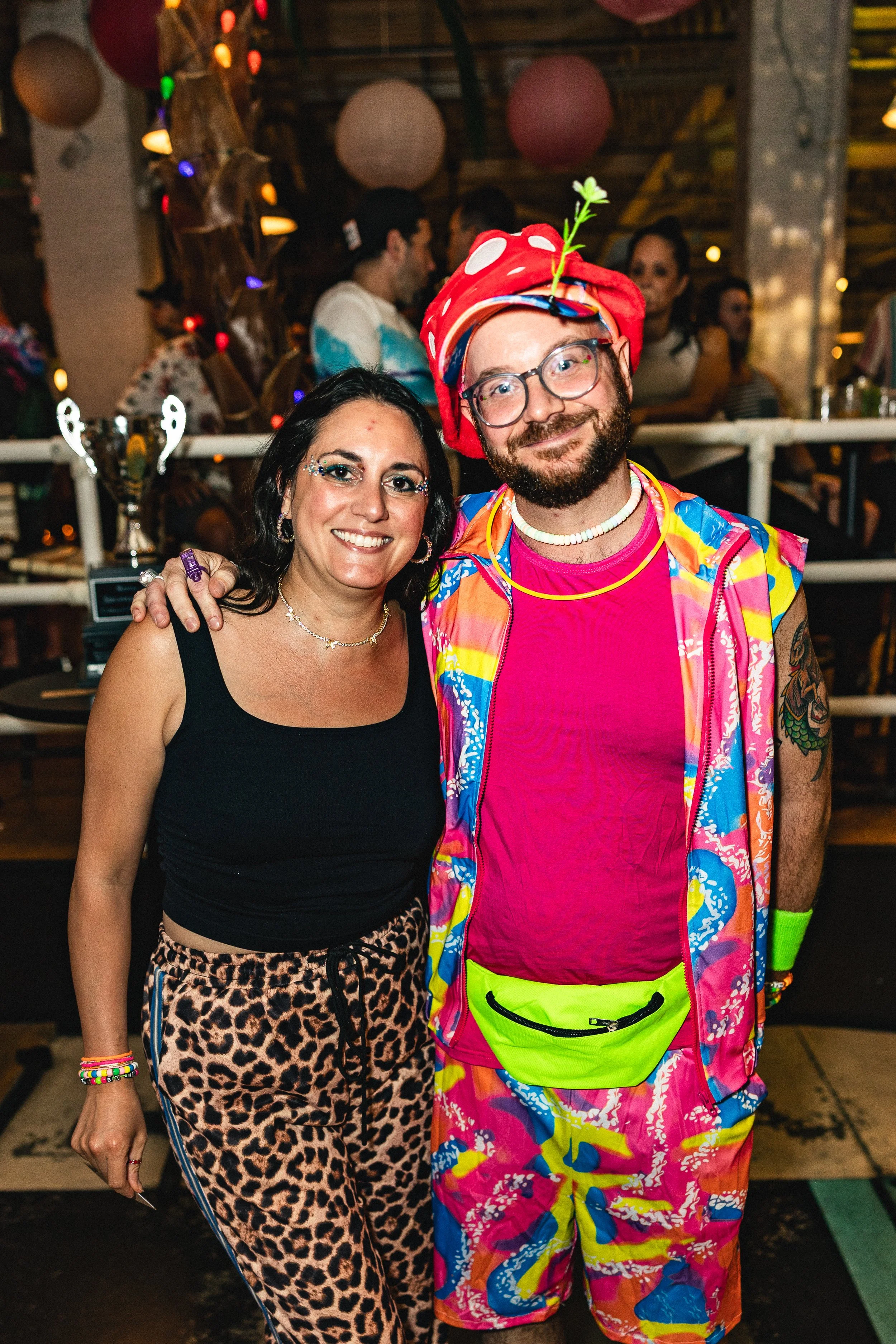 A woman and a man dressed in colorful, playful clothing at a party, smiling at the camera. The woman wears a black sleeveless top and leopard print pants, while the man wears bright, patterned shorts, a pink shirt, and a whimsical hat with a sprout o