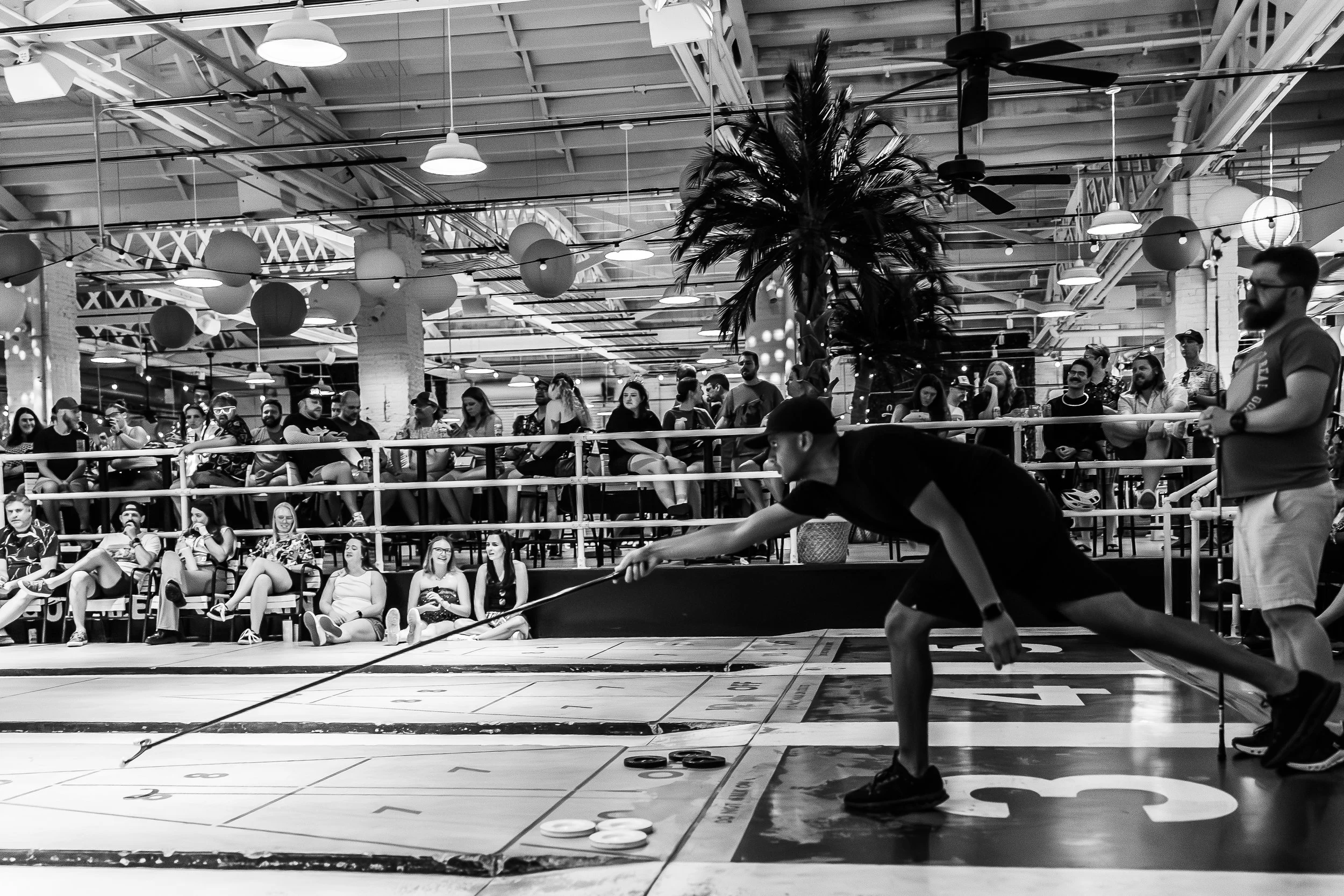 A man playing shuffleboard in front of an audience seated behind a railing, indoor space with hanging lamps, ceiling fans, and decorative trees.