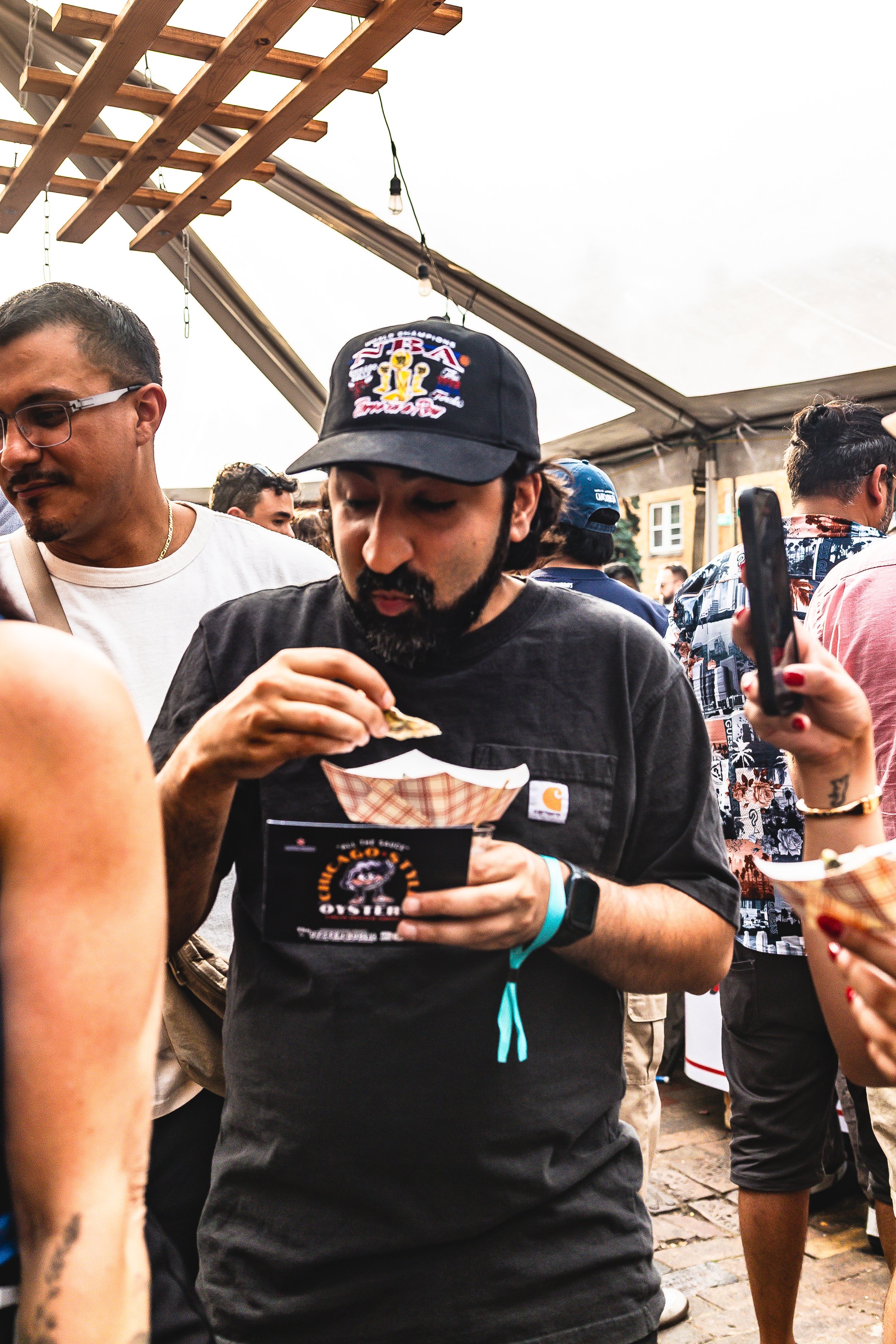 A man wearing a black Carhartt t-shirt and a baseball cap eating food at an outdoor event under a tent with string lights, surrounded by other people