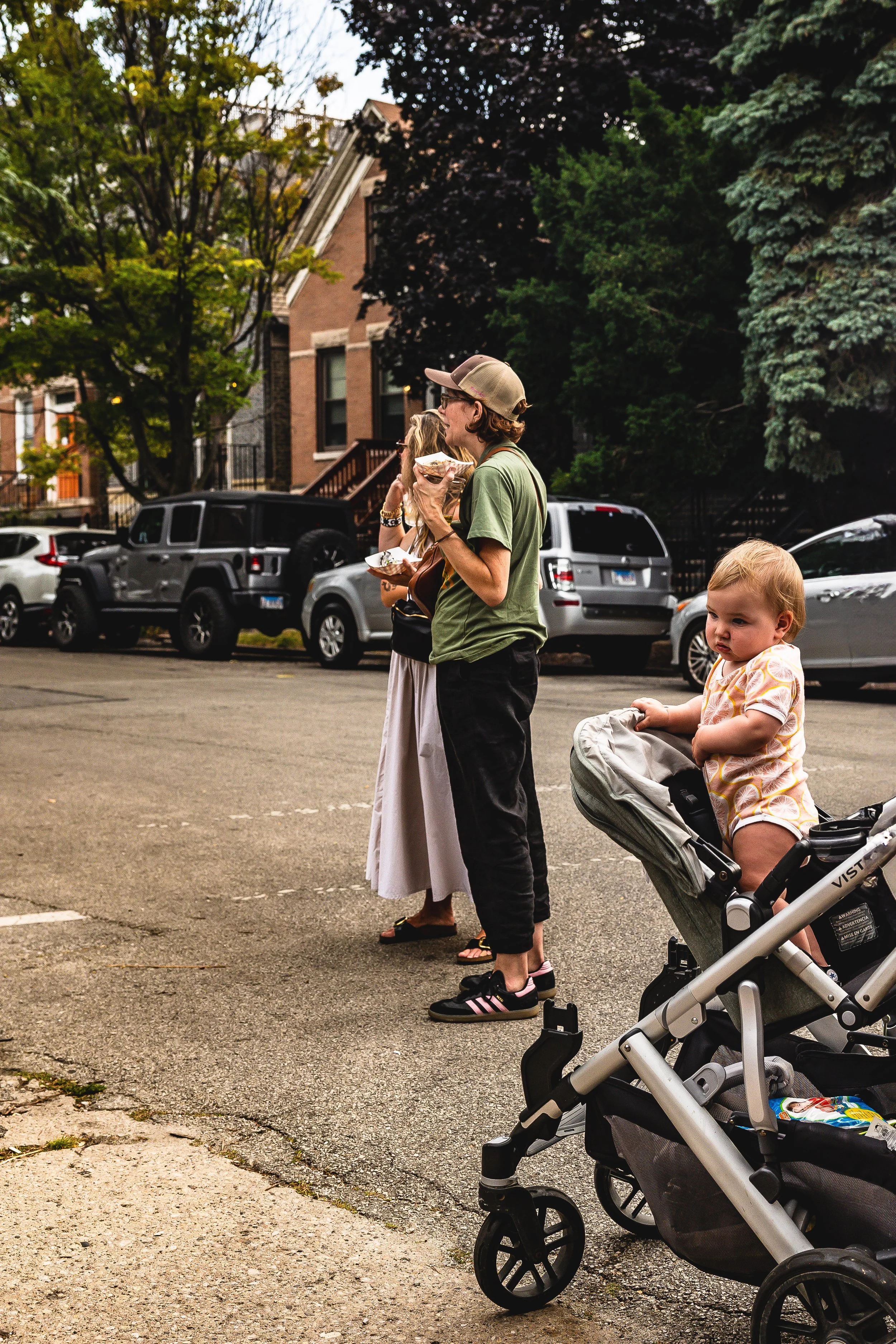 Three people standing by parked cars in a residential area; a woman and a man eating food, and a young child in a stroller.