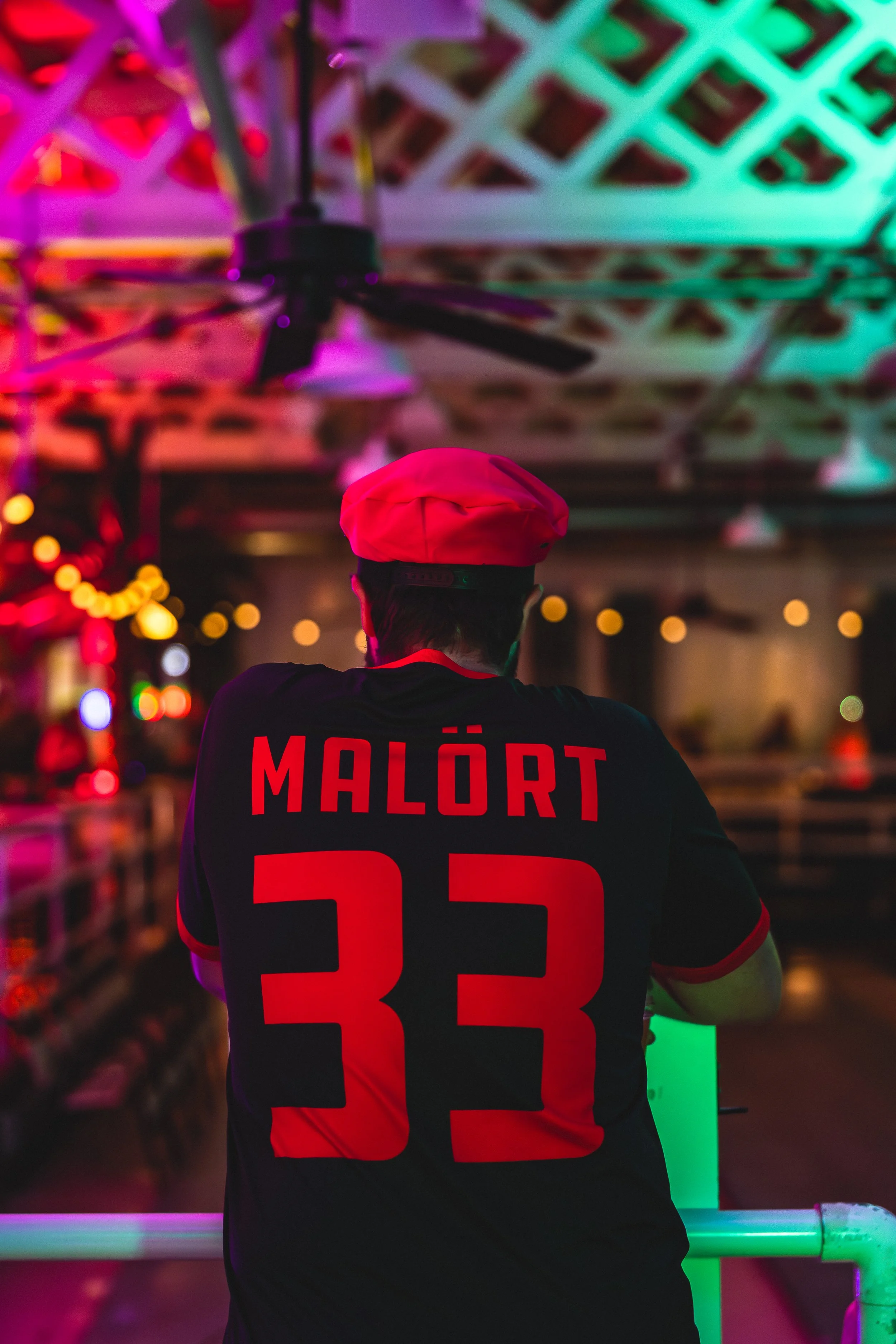 Back of a person wearing a black and red shirt with the name 'Malört' and the number 33, and a red chef hat, standing in a dimly lit, colorful indoor space with purple, green, and orange lighting.