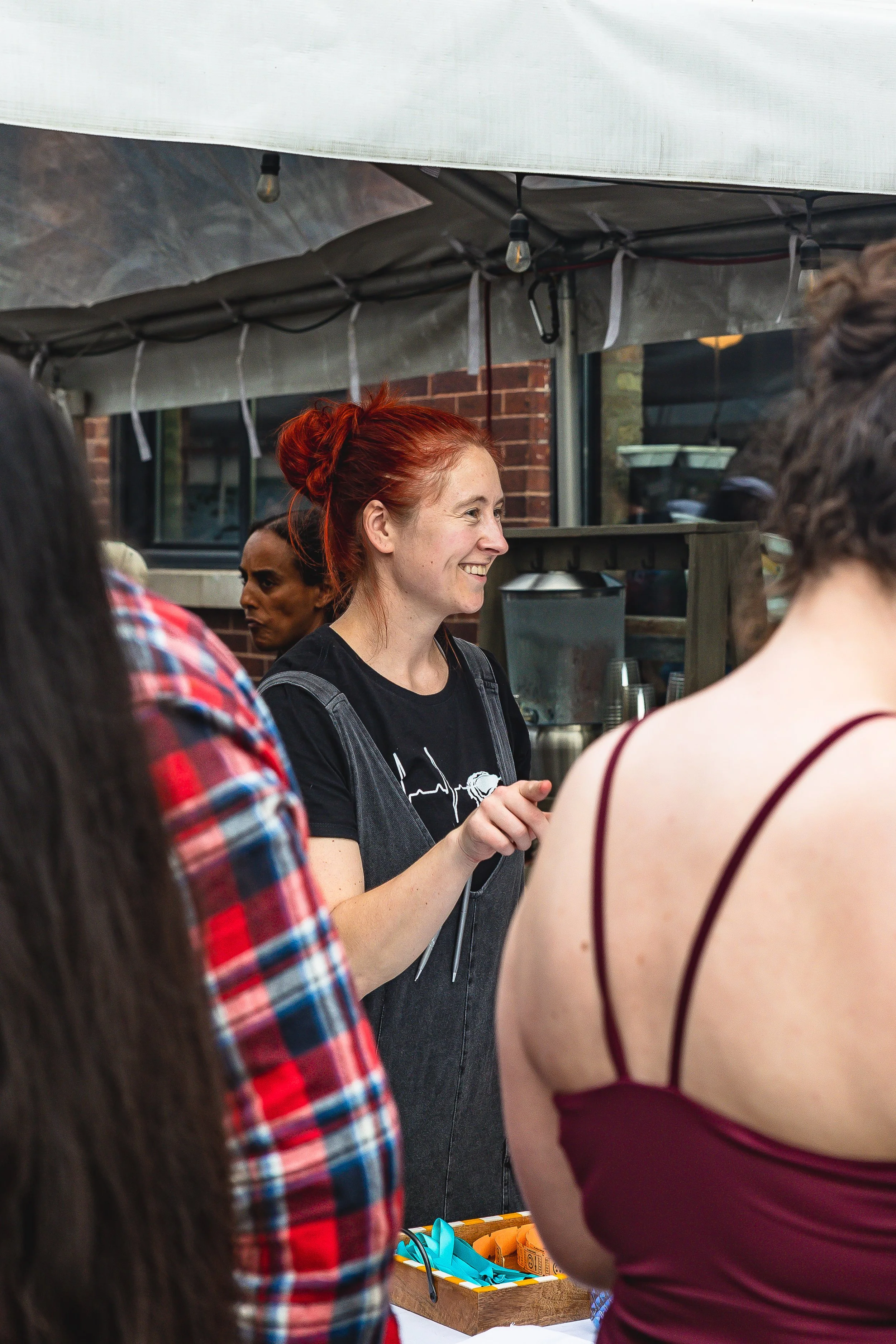 A woman with red hair smiling and talking to a crowd at an outdoor market stall.