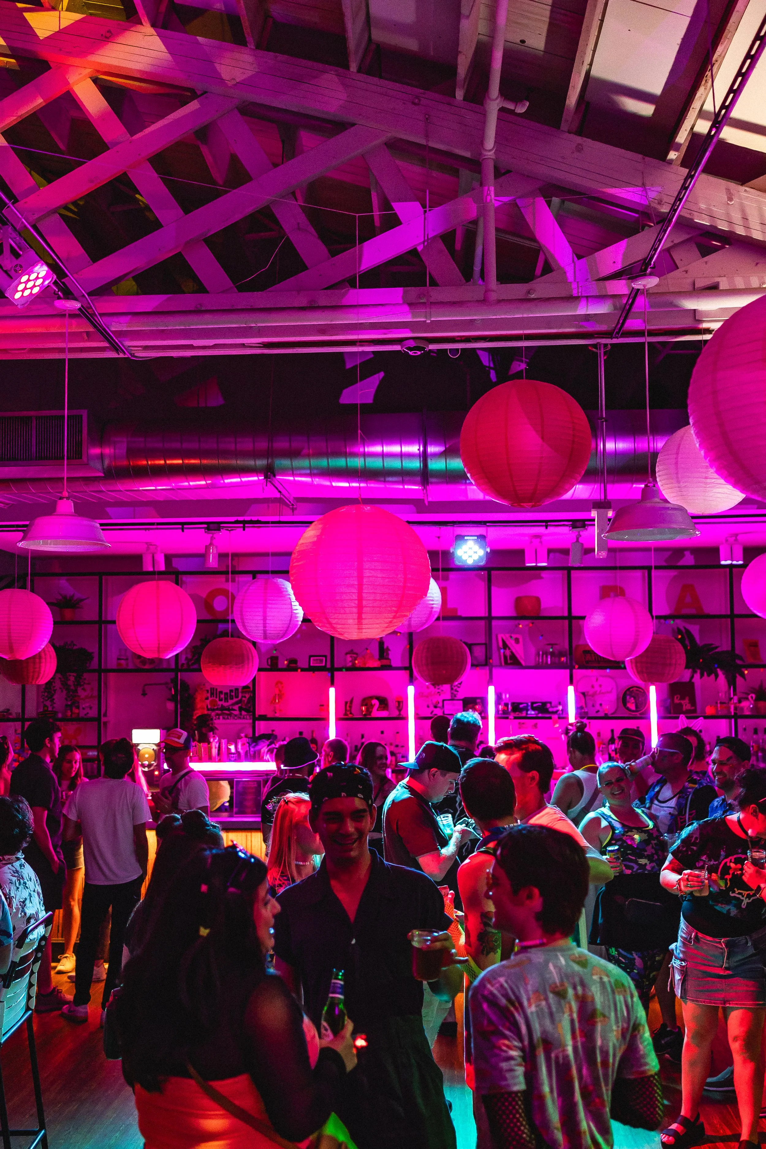 A lively indoor party or club scene with vibrant pink and purple lighting, hanging paper lanterns, and a crowd of people socializing and drinking.