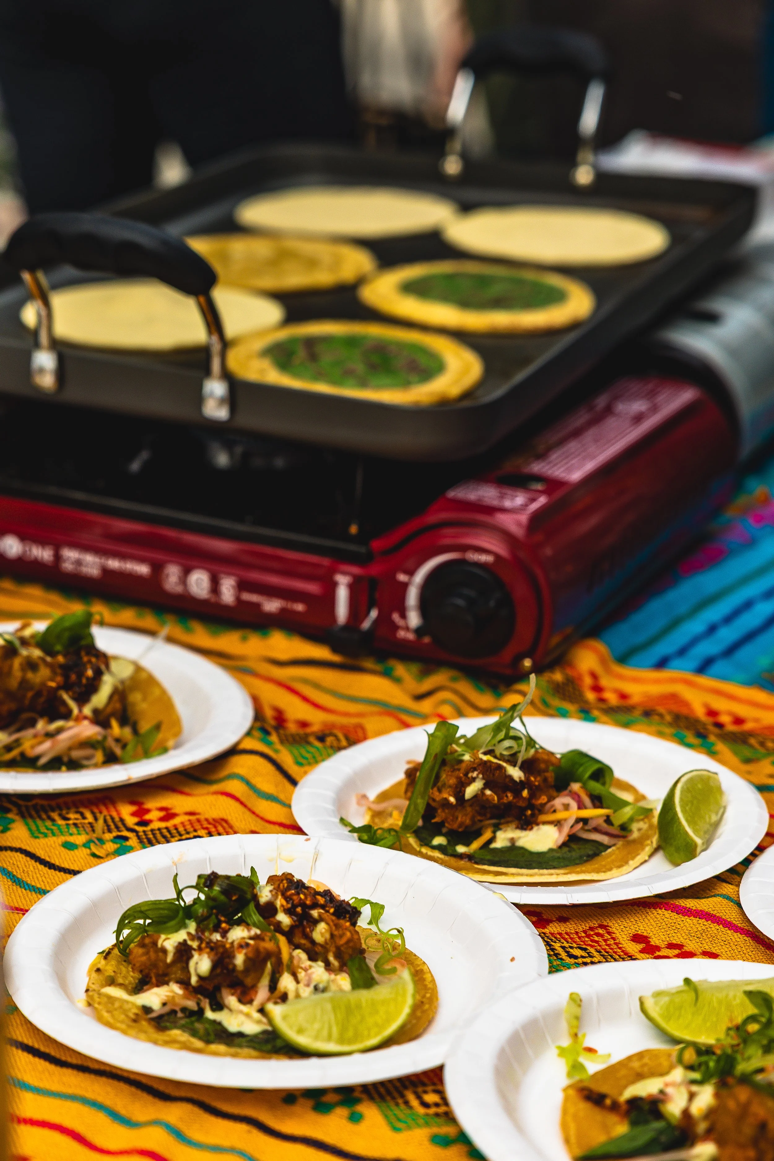 Street food preparation with small plates of tacos garnished with lime wedges and green herbs, cooking on a portable griddle.