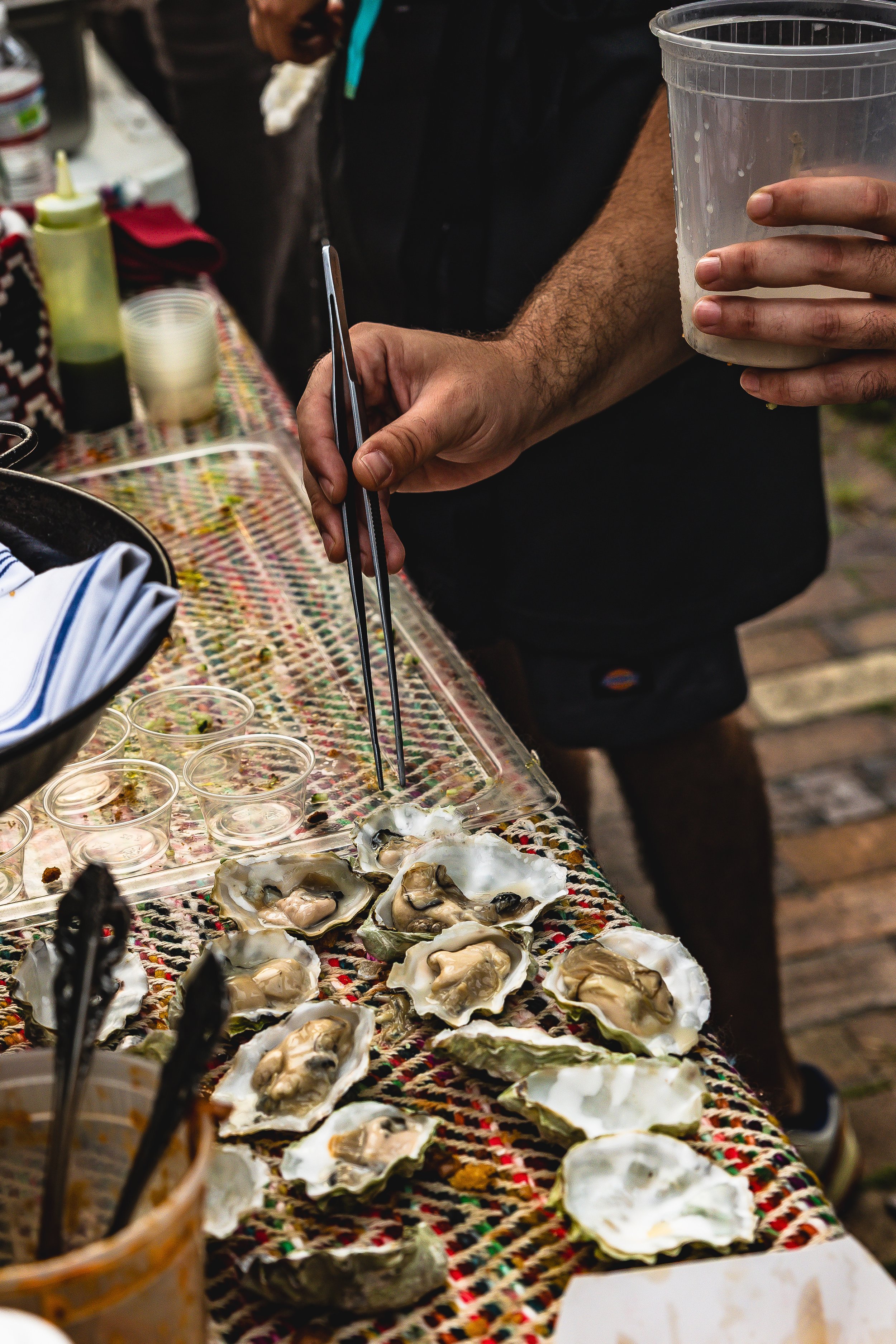A person using tongs to pick up an oyster from a table filled with opened oysters and small empty cups, with a hand holding a plastic container and various condiments and tools in the background.