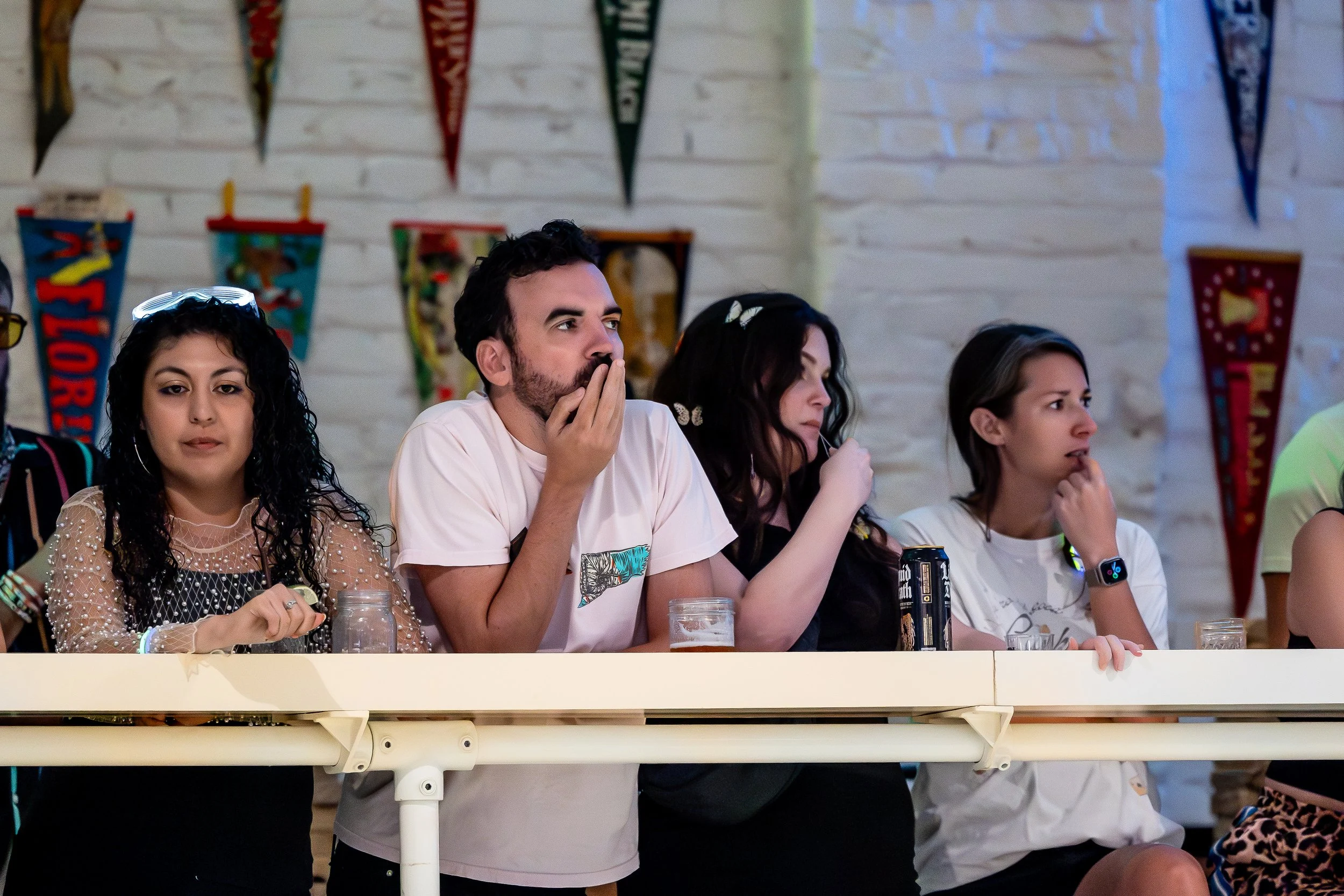 Group of four young adults sitting at a table, watching something with focused and surprised expressions. There are colorful flags hanging on a white brick wall behind them.