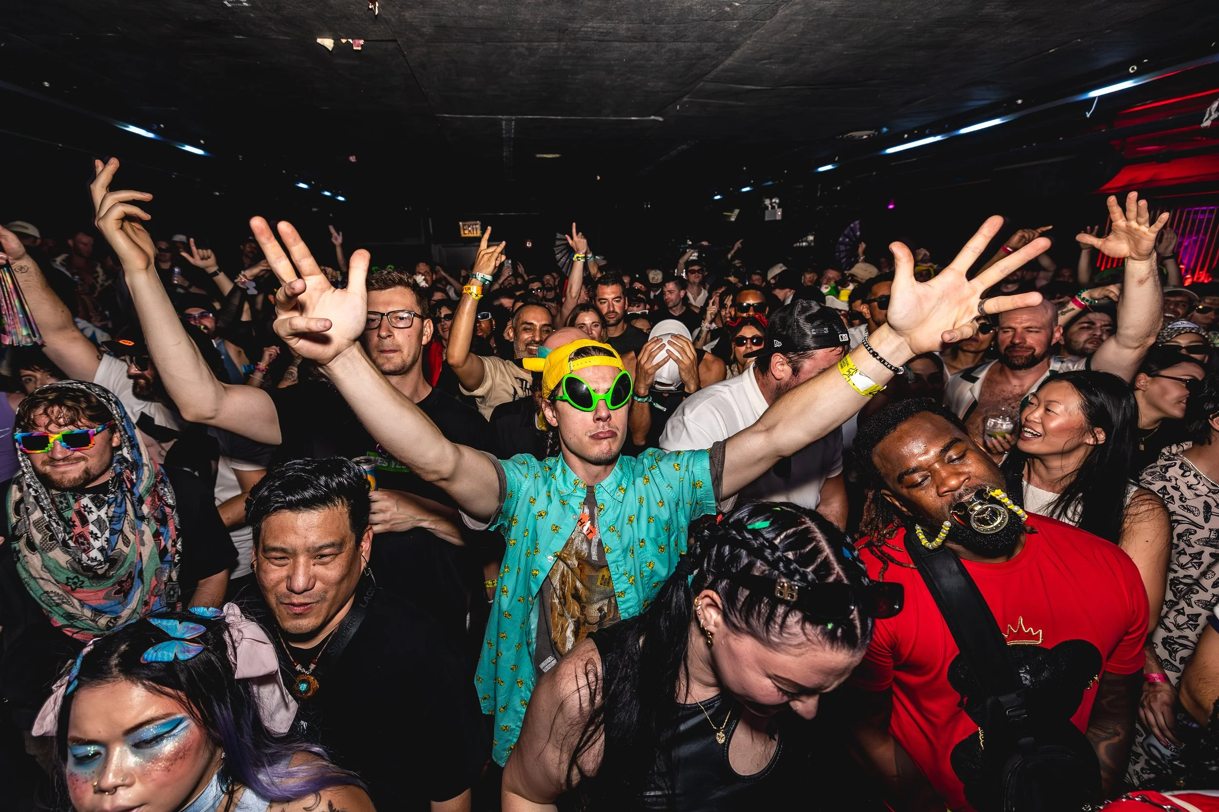 People dancing at a crowded party or concert, wearing colorful and eclectic outfits, with some wearing sunglasses and accessories, in a lively indoor setting.