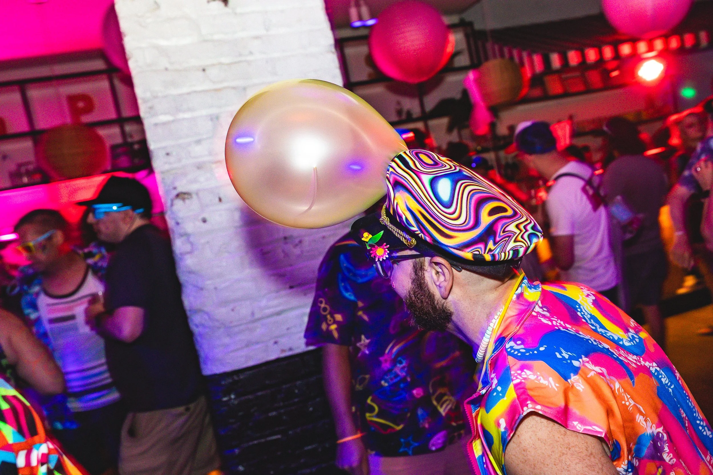 Person wearing a colorful hat and shirt with a large pearl balloon on their head at a lively party with neon lights and other attendees.