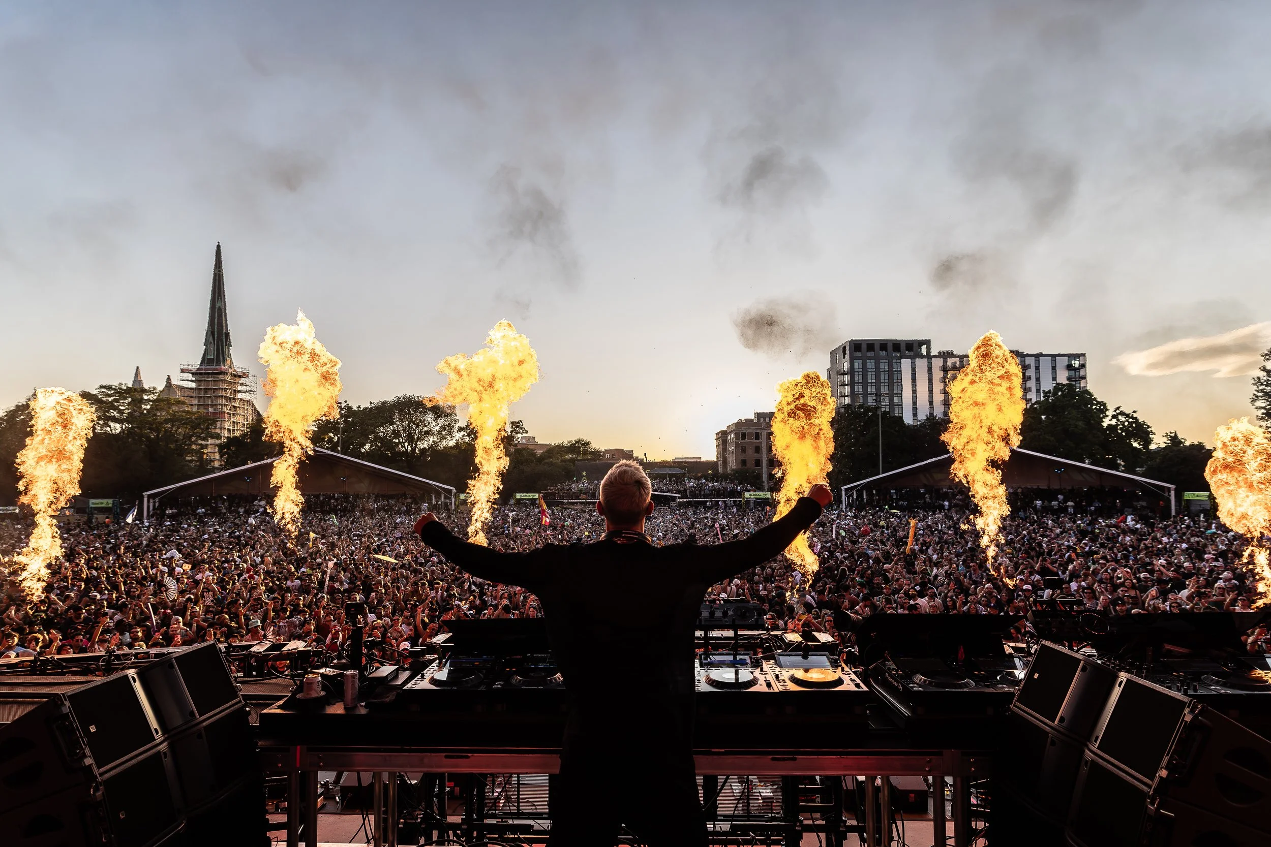 A DJ performs on stage at an outdoor music festival with pyrotechnics, a large crowd, and city buildings in the background.