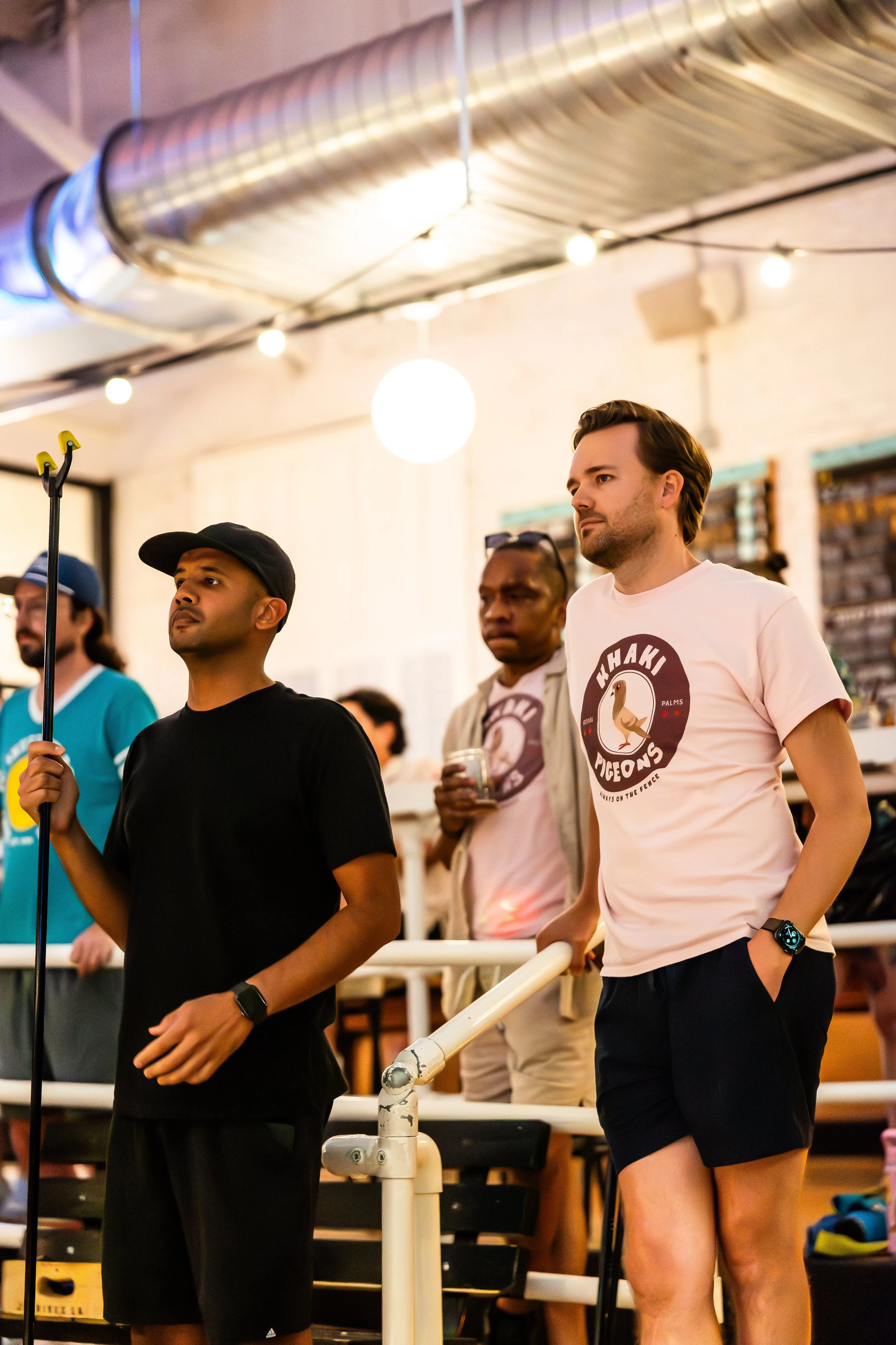 A group of diverse men standing indoors under string lights and a large air duct, paying attention to something out of frame.