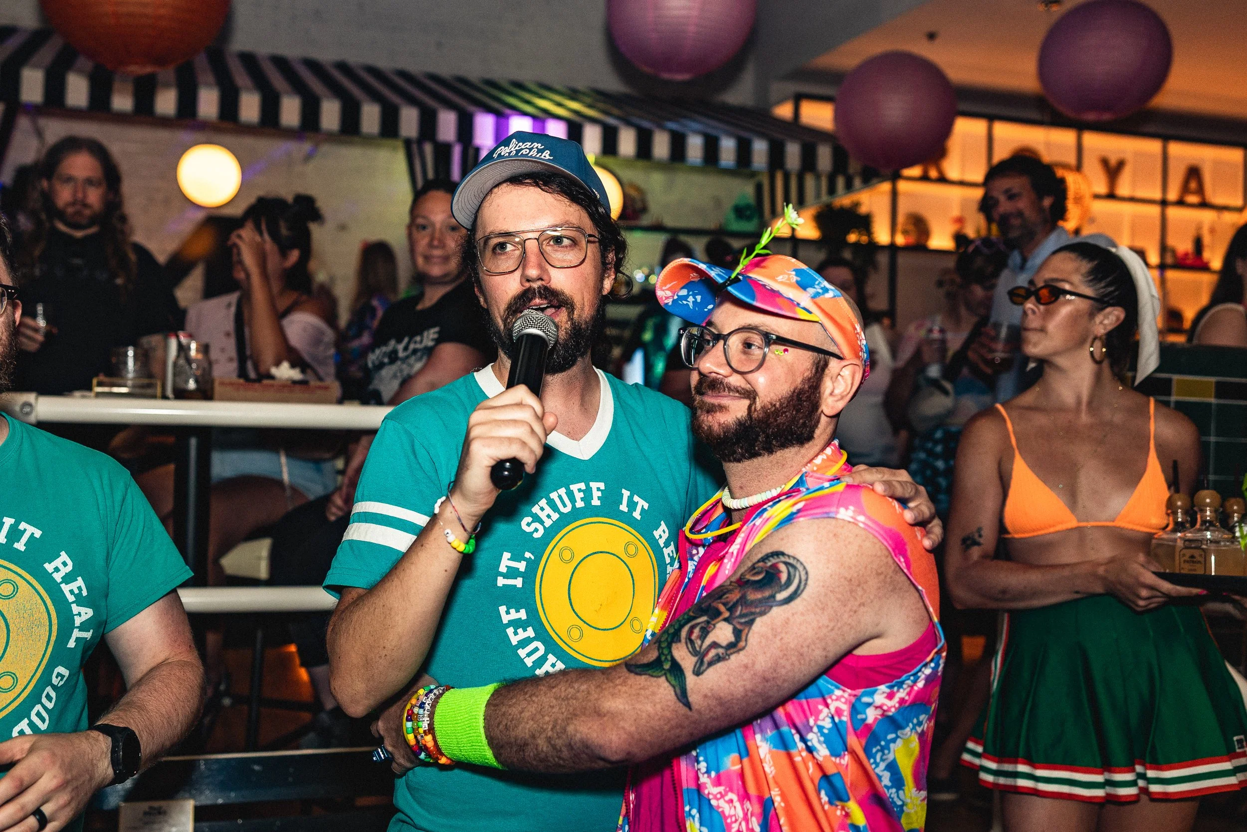 A man with glasses and a colorful hat, being hugged by another man in a turquoise T-shirt, at a lively party with daytime themed decorations and people in casual and colorful outfits.