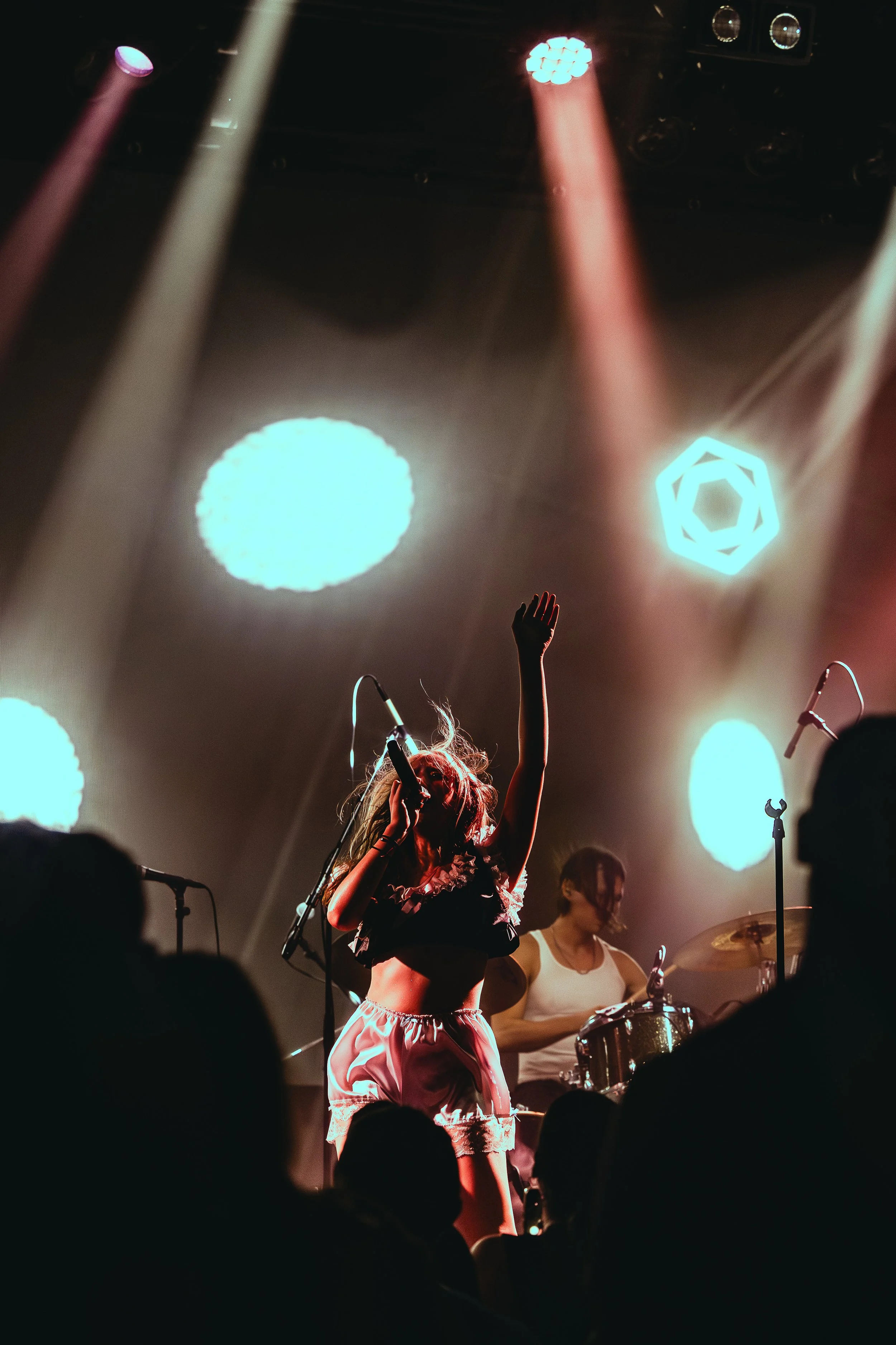 A female singer with long flowing hair singing into a microphone on stage, raising her hand, with a drummer playing in the background, concert stage with colorful lights and audience silhouettes in the foreground.