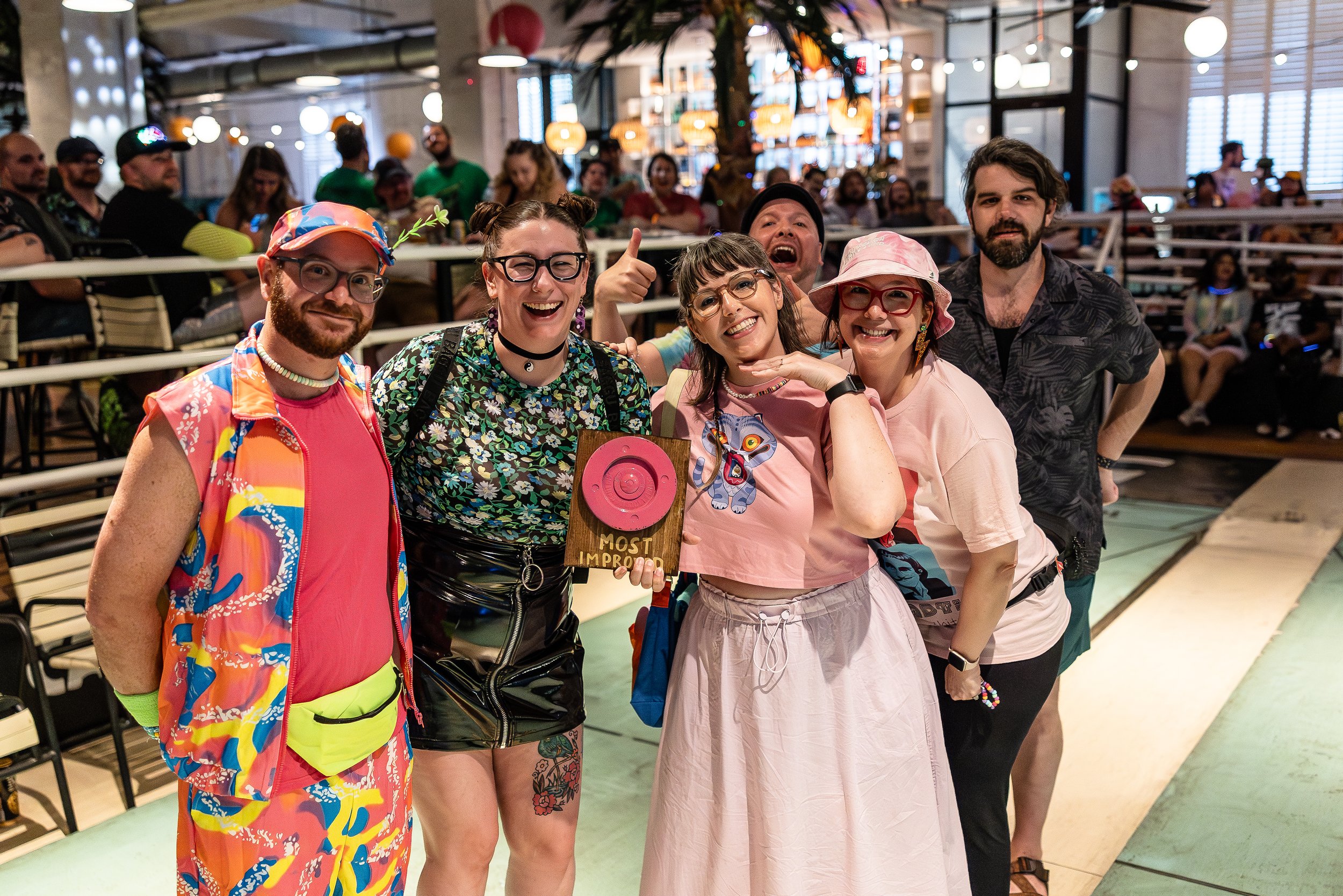 Group of six friends smiling and posing at an indoor event or party, with people in the background and colorful decorations.