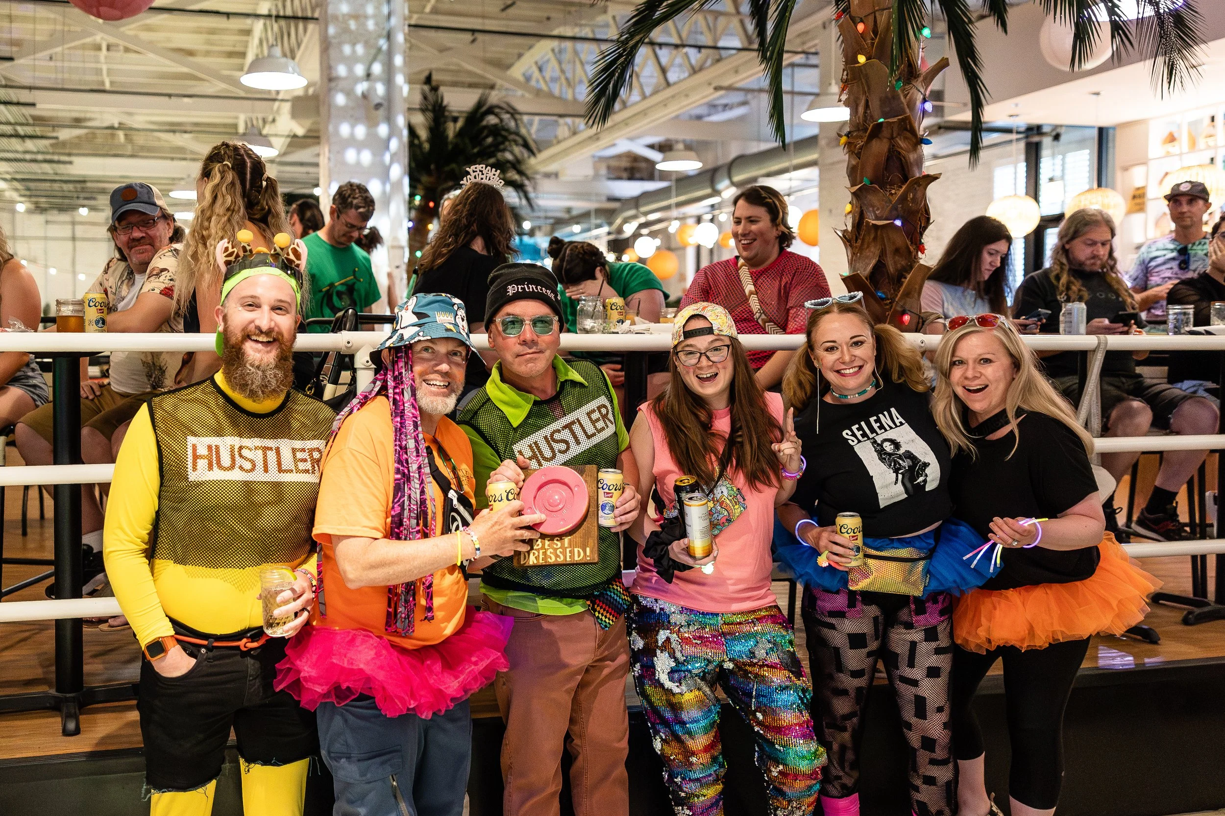 Group of people celebrating at a party, wearing colorful and playful outfits, some with 'HUSTLER' printed shirts, and others in bright skirts, with a decorated indoor setting.