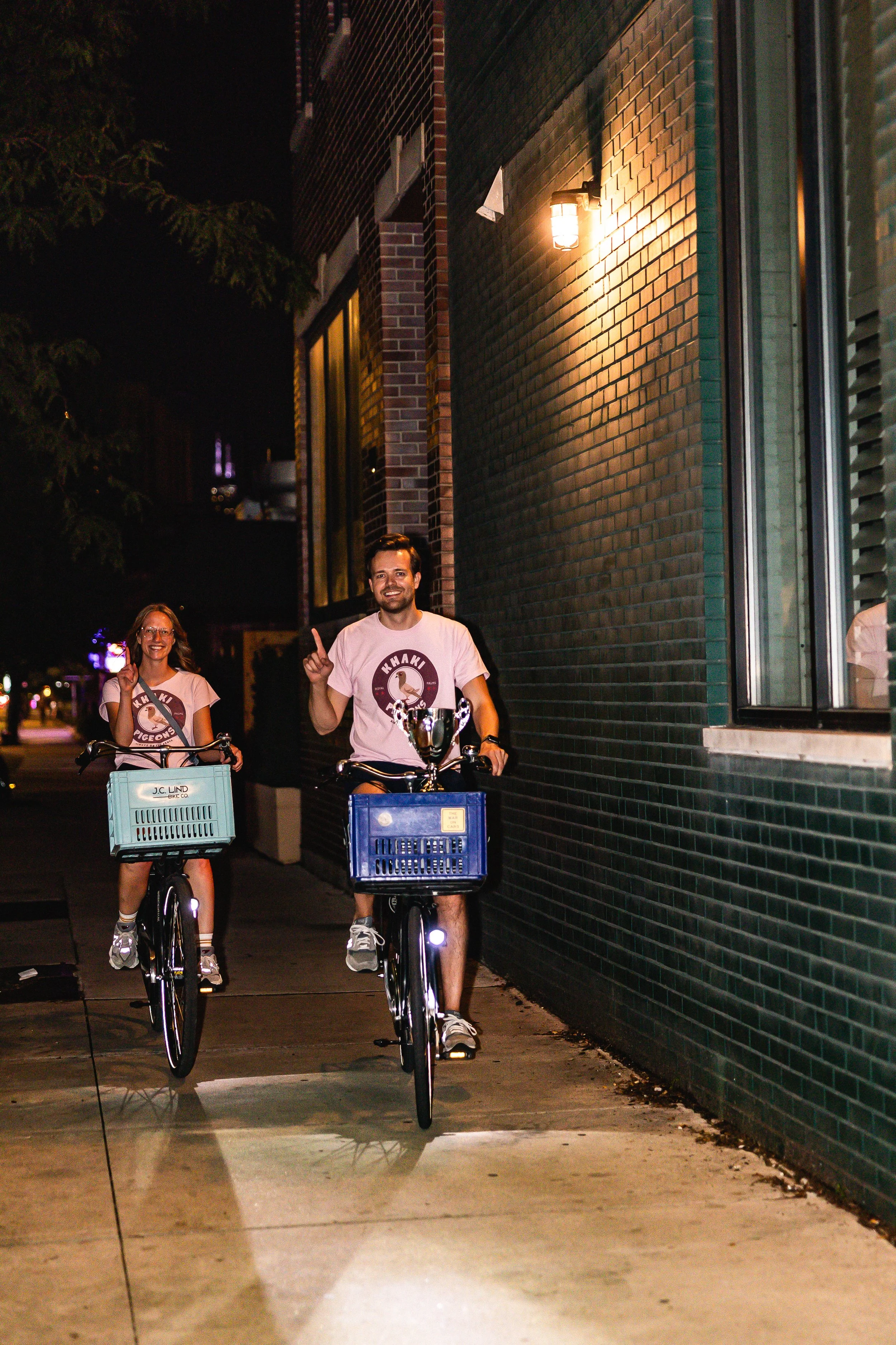 Two people riding bicycles at night on a city sidewalk, smiling, with one person raising one finger, wearing matching T-shirts with a logo, with a brick building and wall-mounted light in the background.