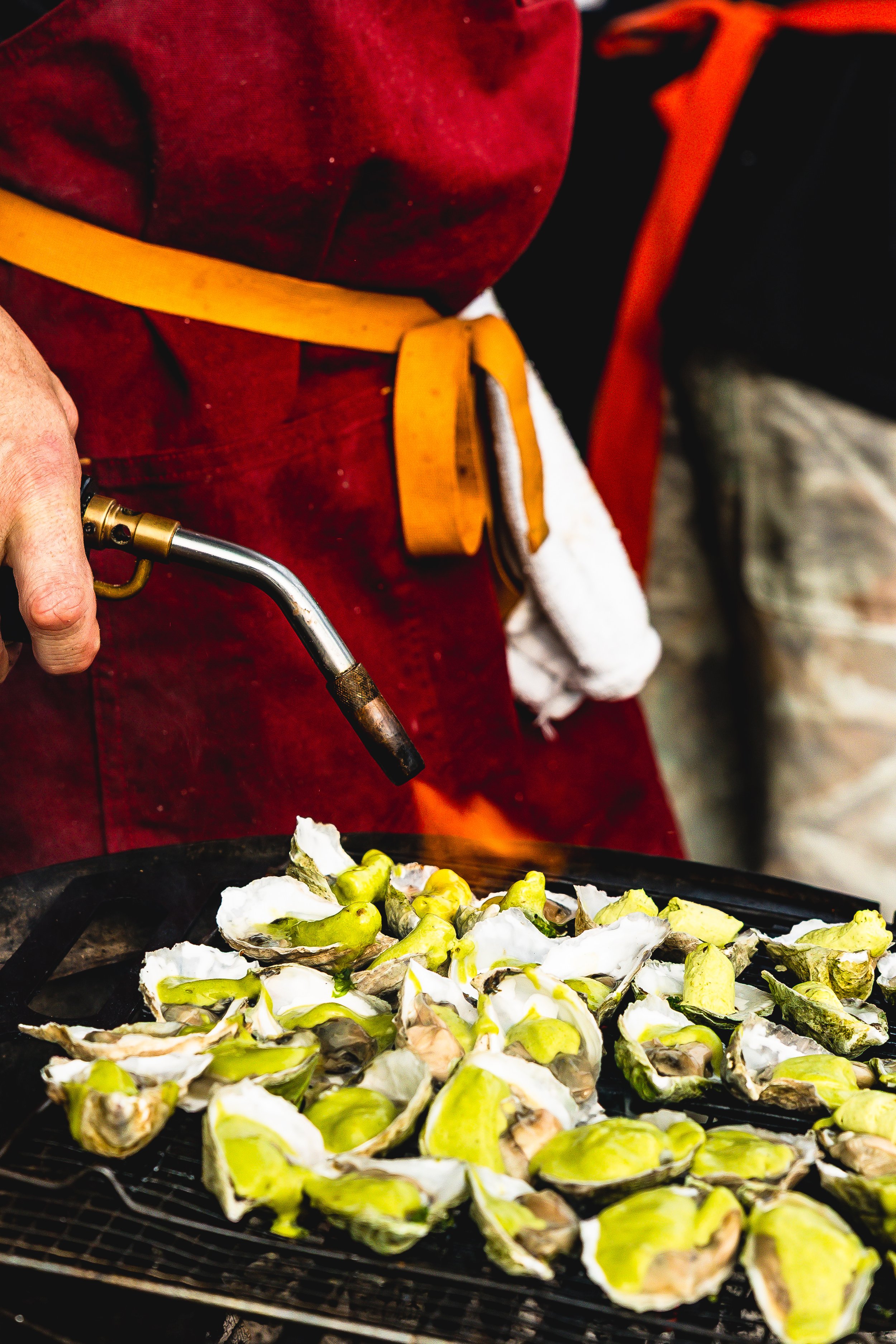 Person using a culinary torch to roast oysters on a grill, wearing a red apron and a yellow waist apron.
