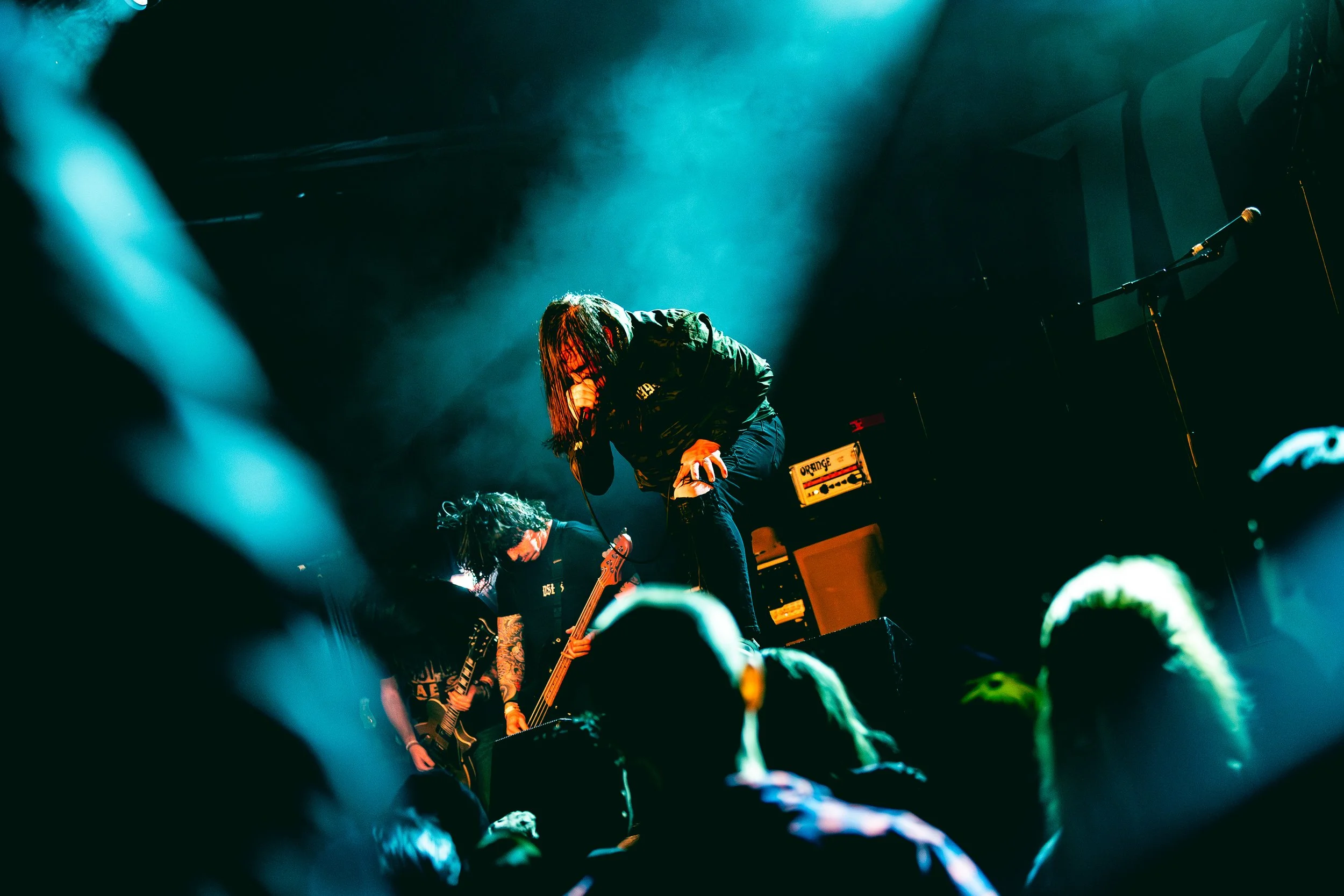 Norma Jean performing live on stage with a lead singer passionately singing into a microphone, and guitarists playing nearby. Audience members are visible in the foreground.