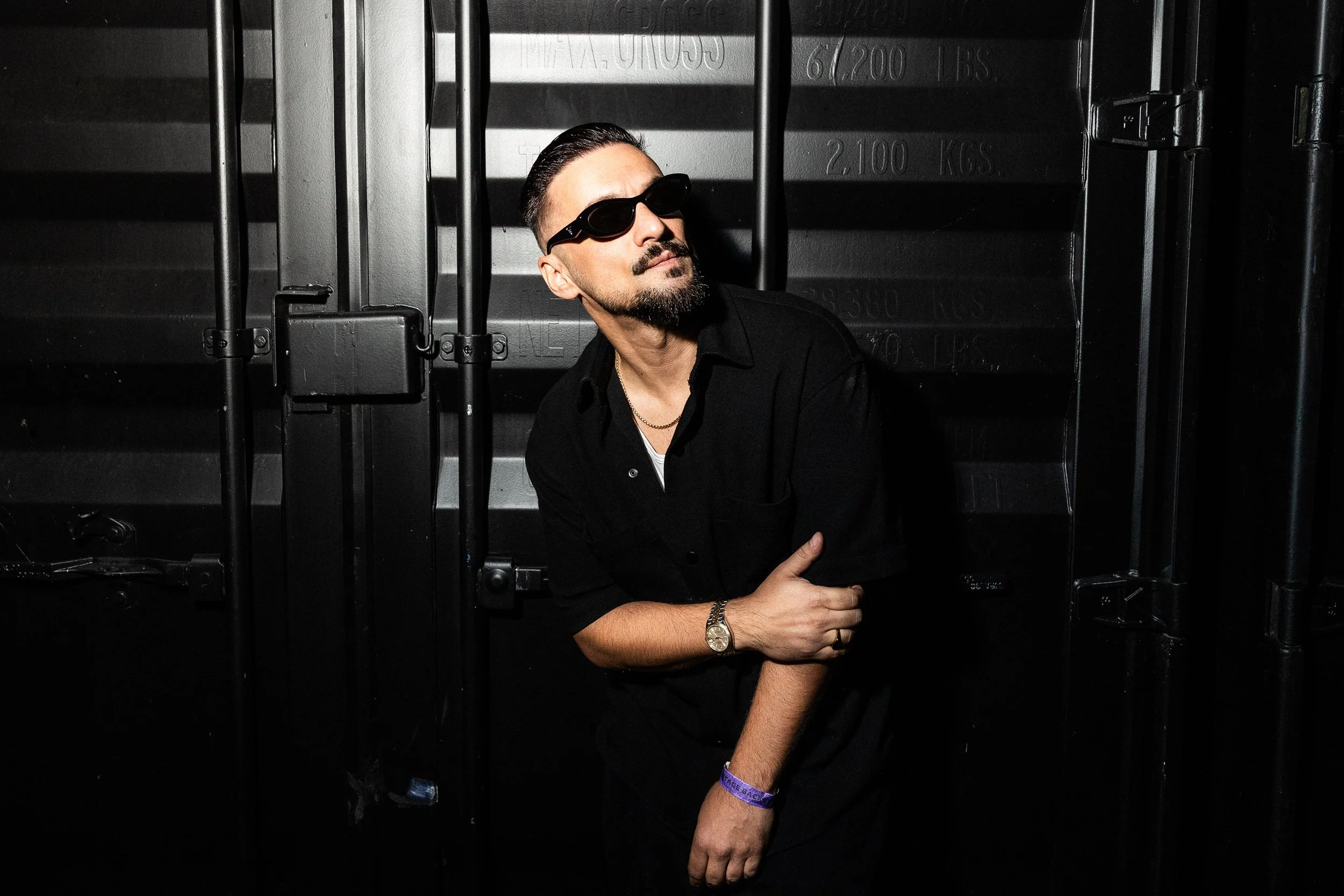 Man with dark sunglasses, beard, and slicked-back hair standing in front of a black metallic wall, wearing a black shirt and a watch.