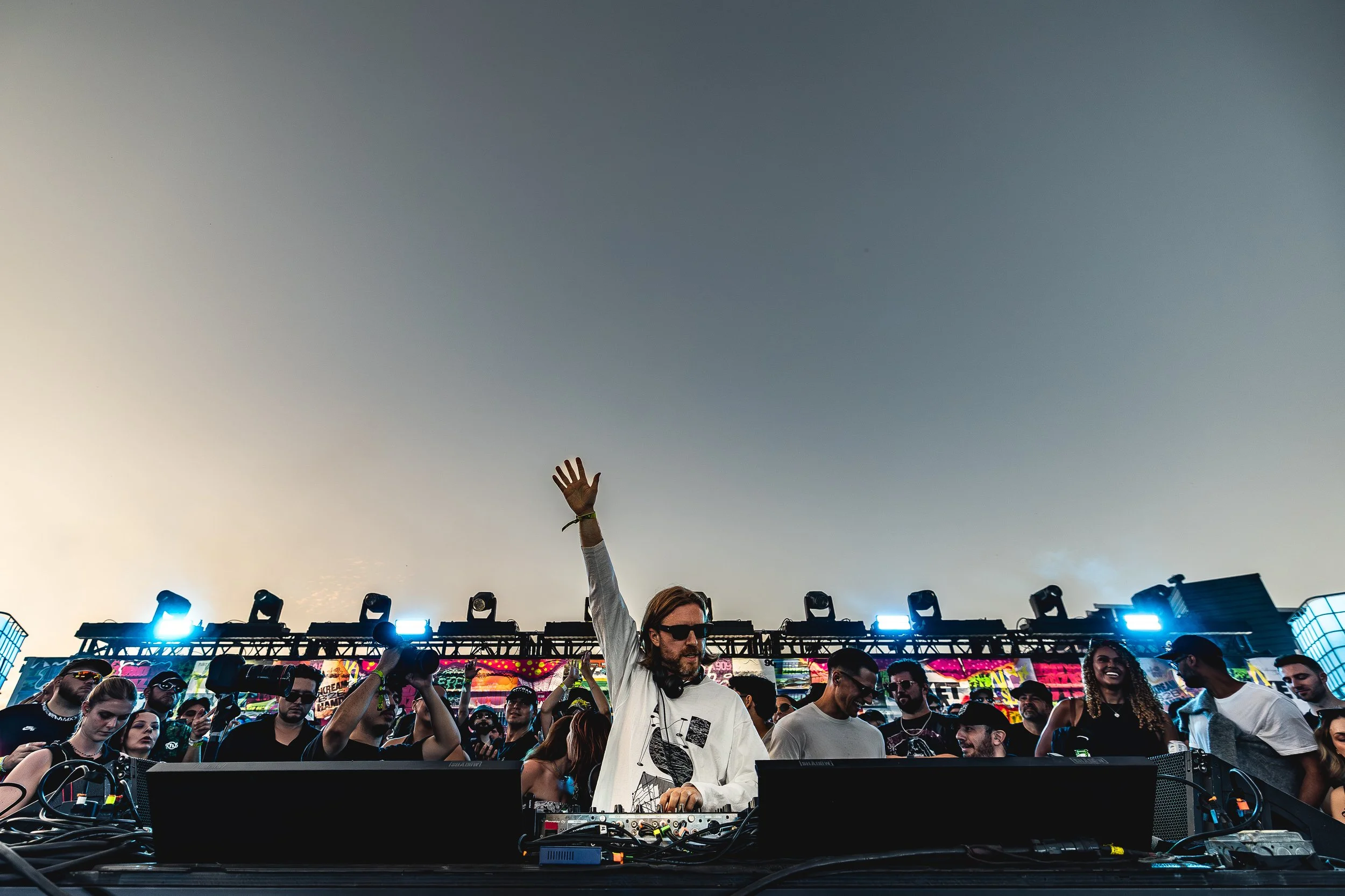 DJ performing at an outdoor music festival during sunset with a crowd of attendees dancing and enjoying the event.