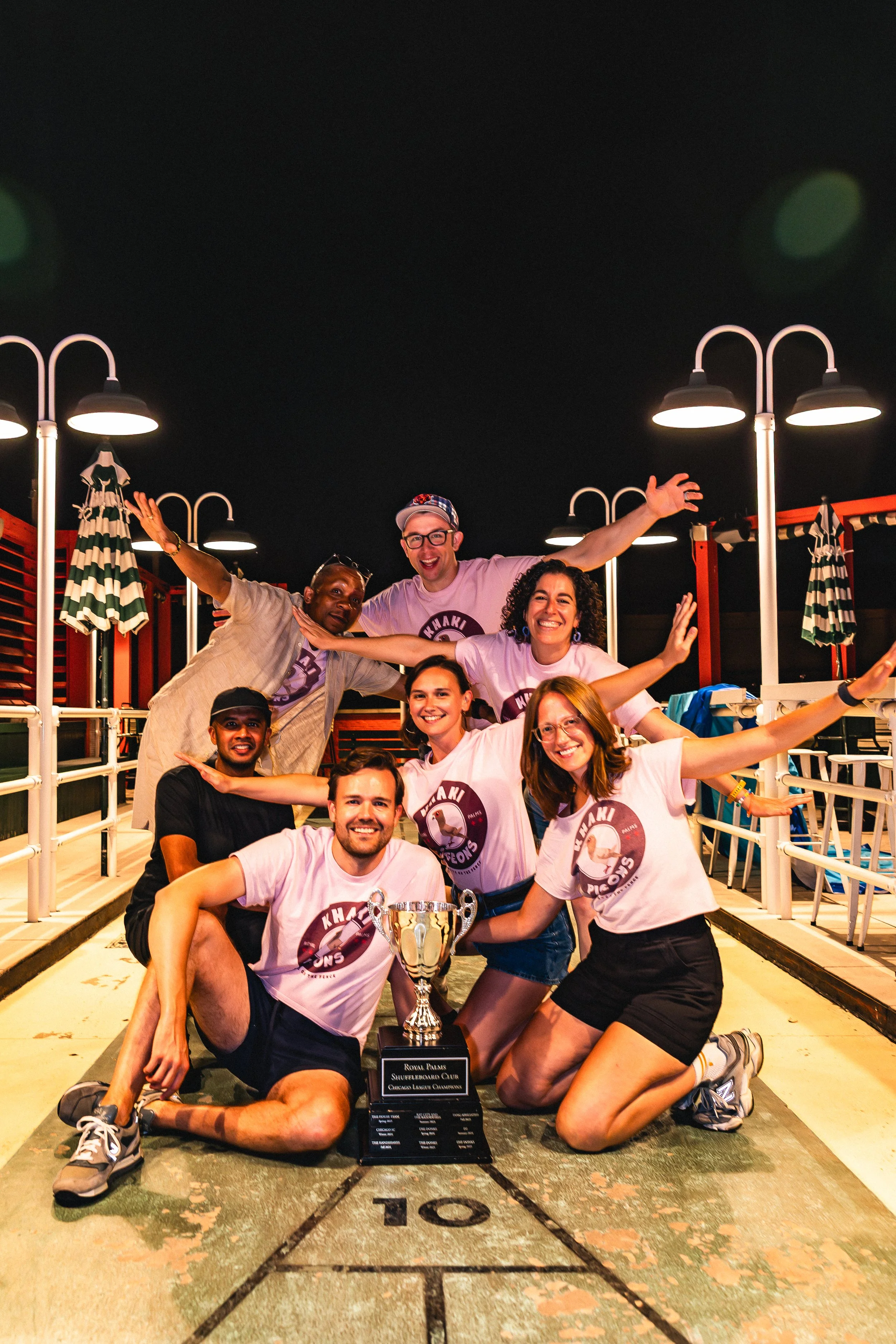Group of people celebrating with a trophy at shuffleboard court at night.