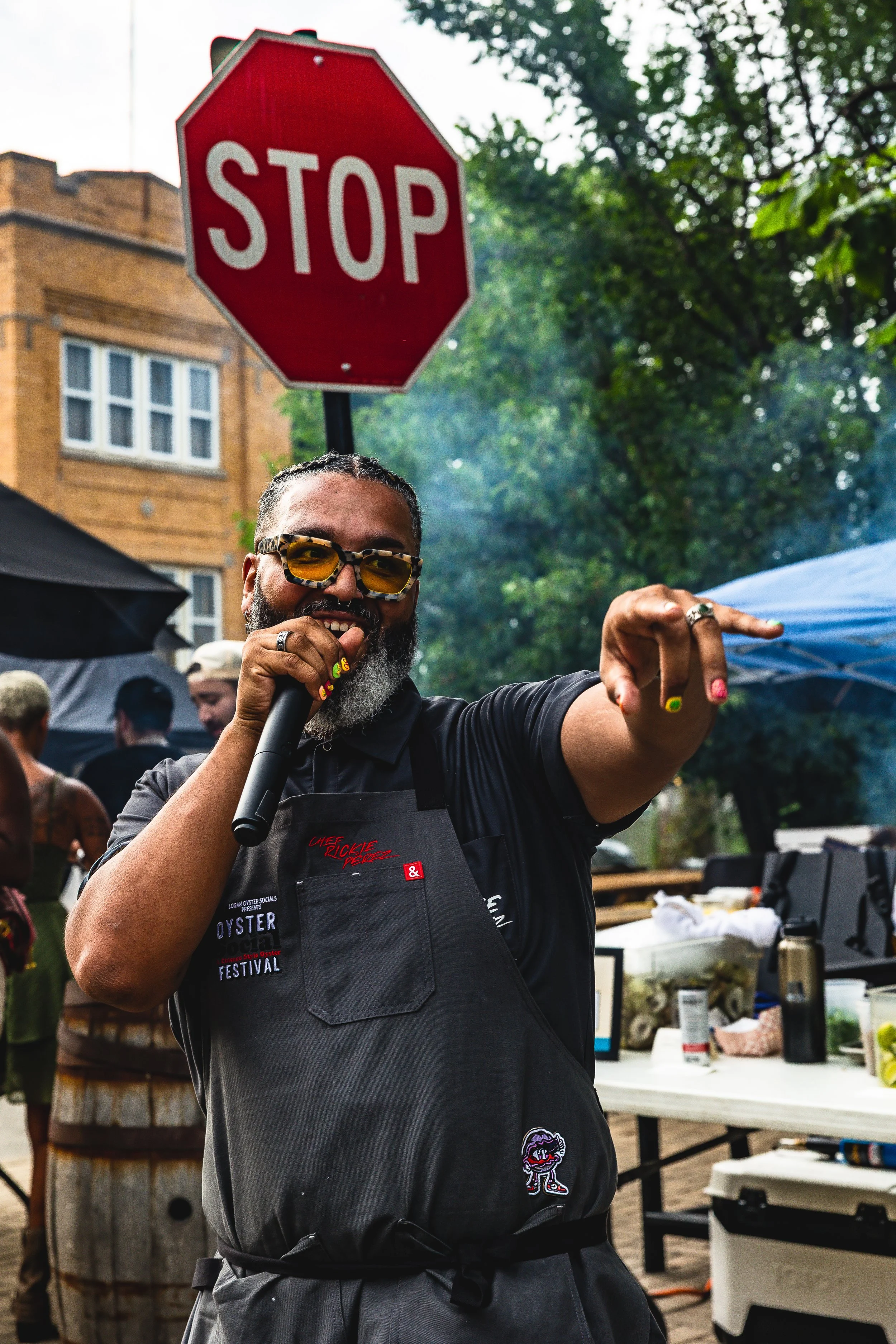 A man with sunglasses and a beard holding a microphone, smiling, pointing forward, standing near a stop sign at an outdoor event with a table and other people in the background.
