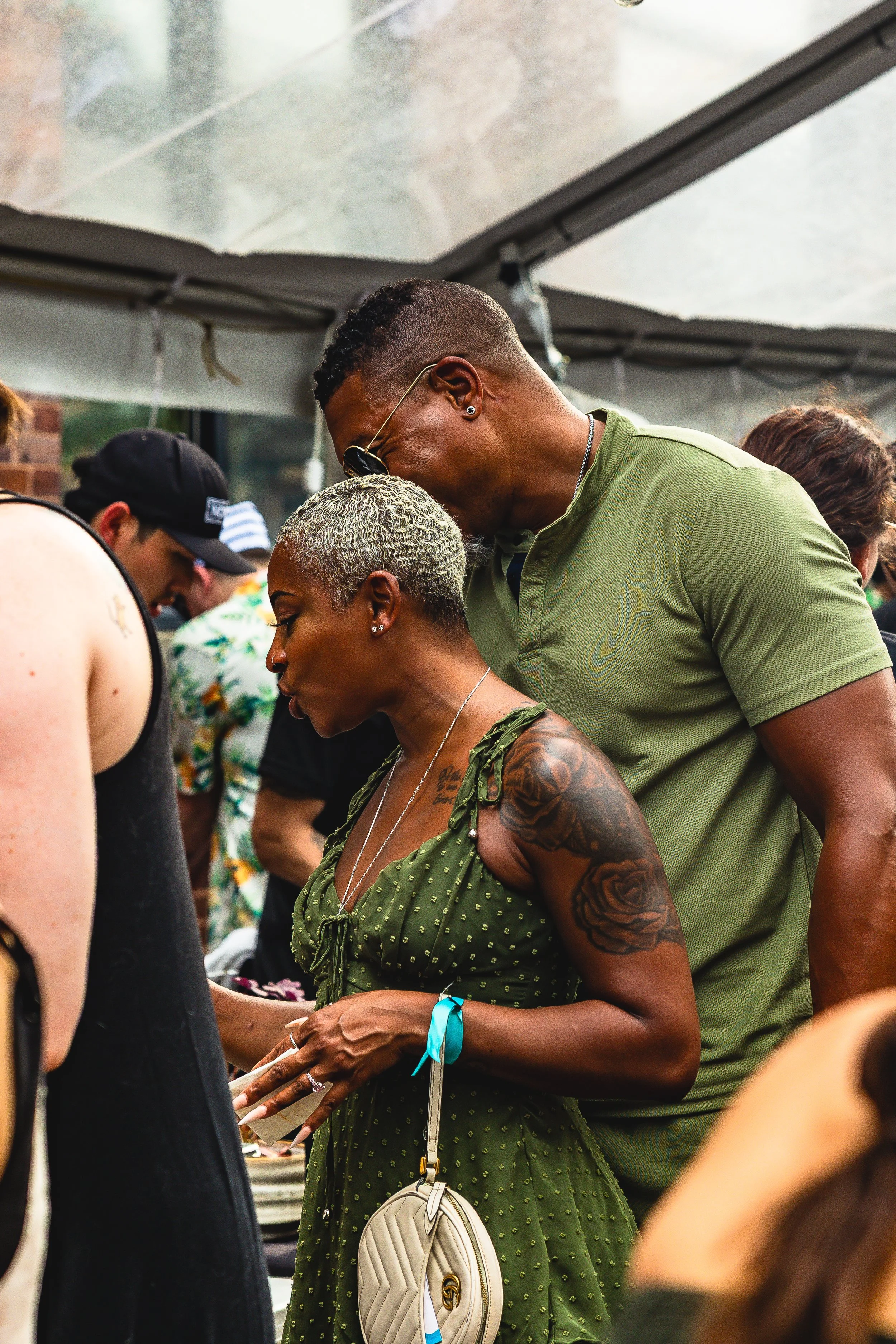 A woman with short, gray curly hair wearing a green dress with small polka dots and a tattoo on her arm, standing in front of a man with dark sunglasses and a green shirt, at an outdoor gathering.