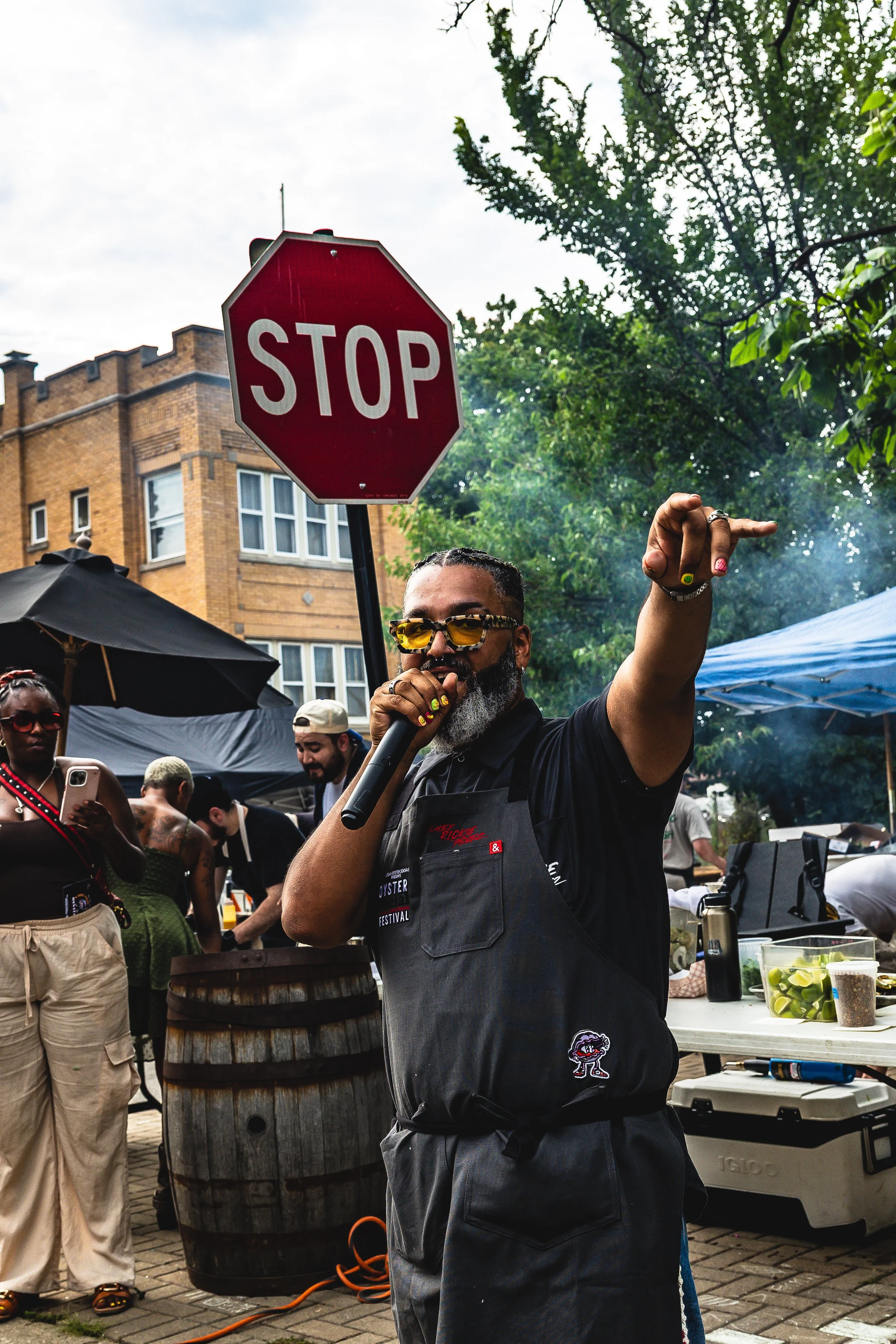 A man in sunglasses speaking into a microphone at an outdoor event, holding a stop sign. People around are taking photos and socializing underneath umbrellas and tents.