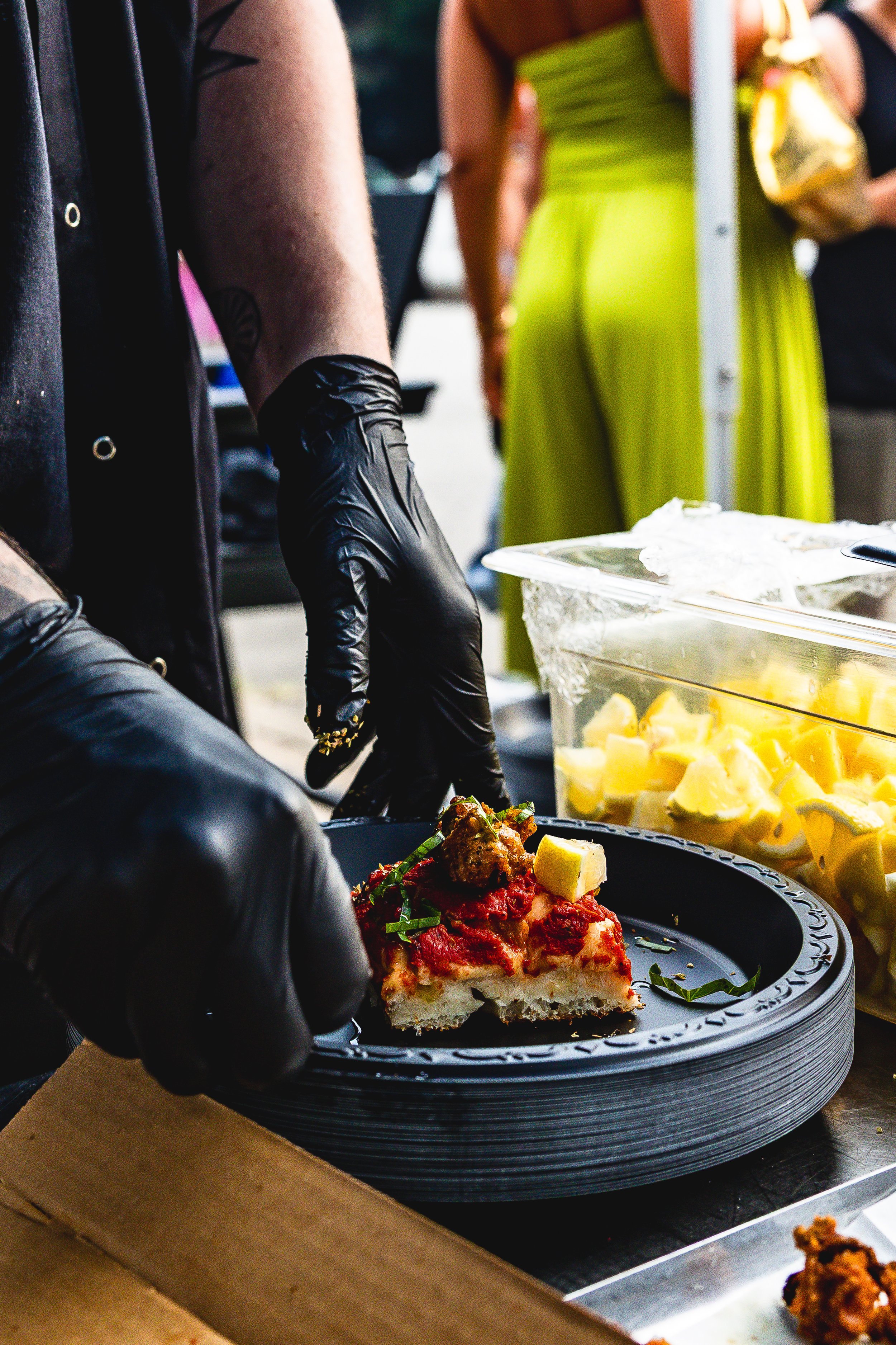 Person wearing black gloves placing a slice of pizza on a black plate at a food event.
