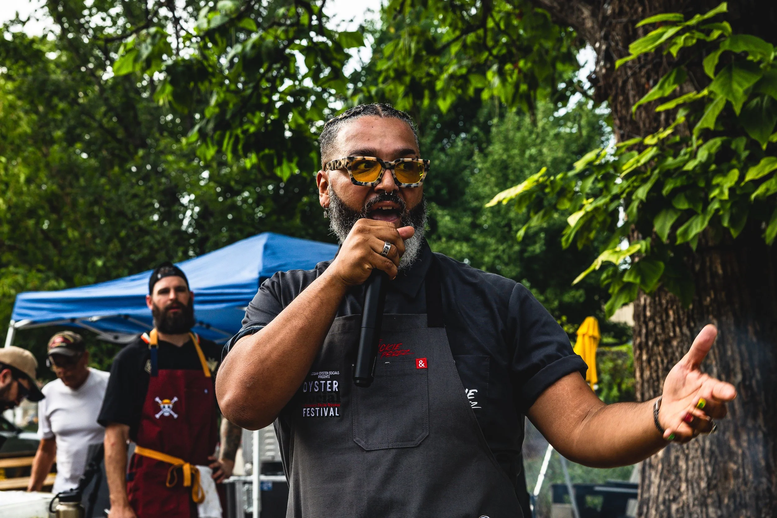 Man speaking into a microphone at an outdoor event, wearing sunglasses and a black apron, with trees and other people in the background