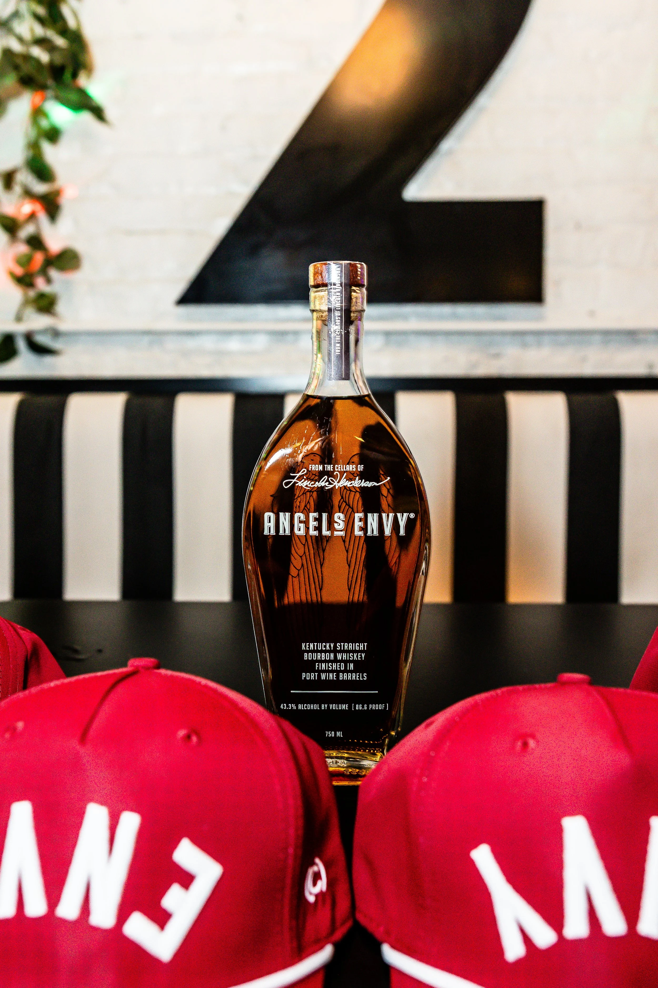 A bottle of Angels Envy Kentucky Straight Bourbon Whiskey on a table, with red caps and white text, in front of a black and white striped background and a large black number 2 on a white wall.