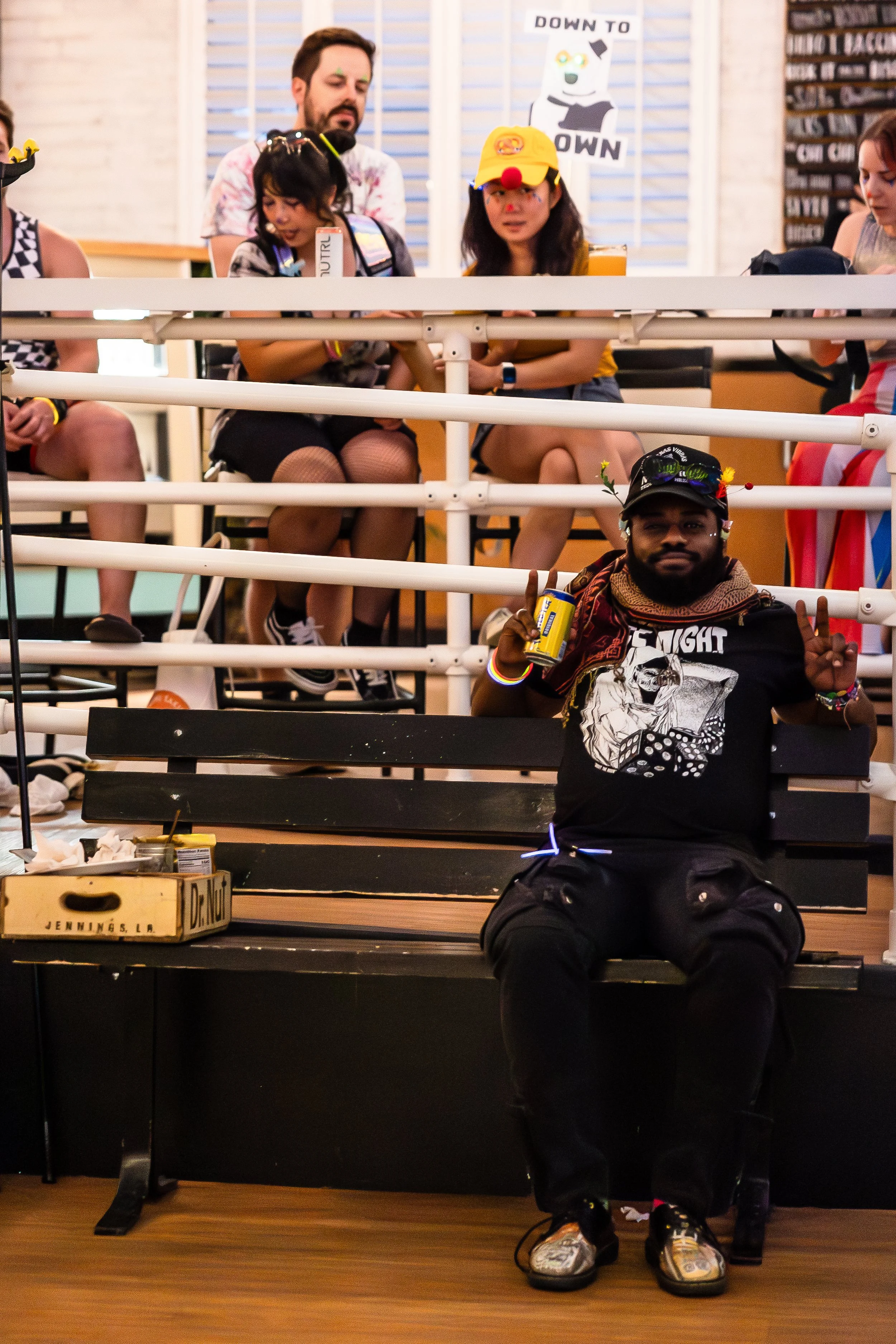 Group of people sitting on bleachers, one person in the front wearing a clown hat and holding a soda, others dressed casually with some wearing costumes like a yellow hat with a red nose. A sign in the background says "DOWN TO BOWN."