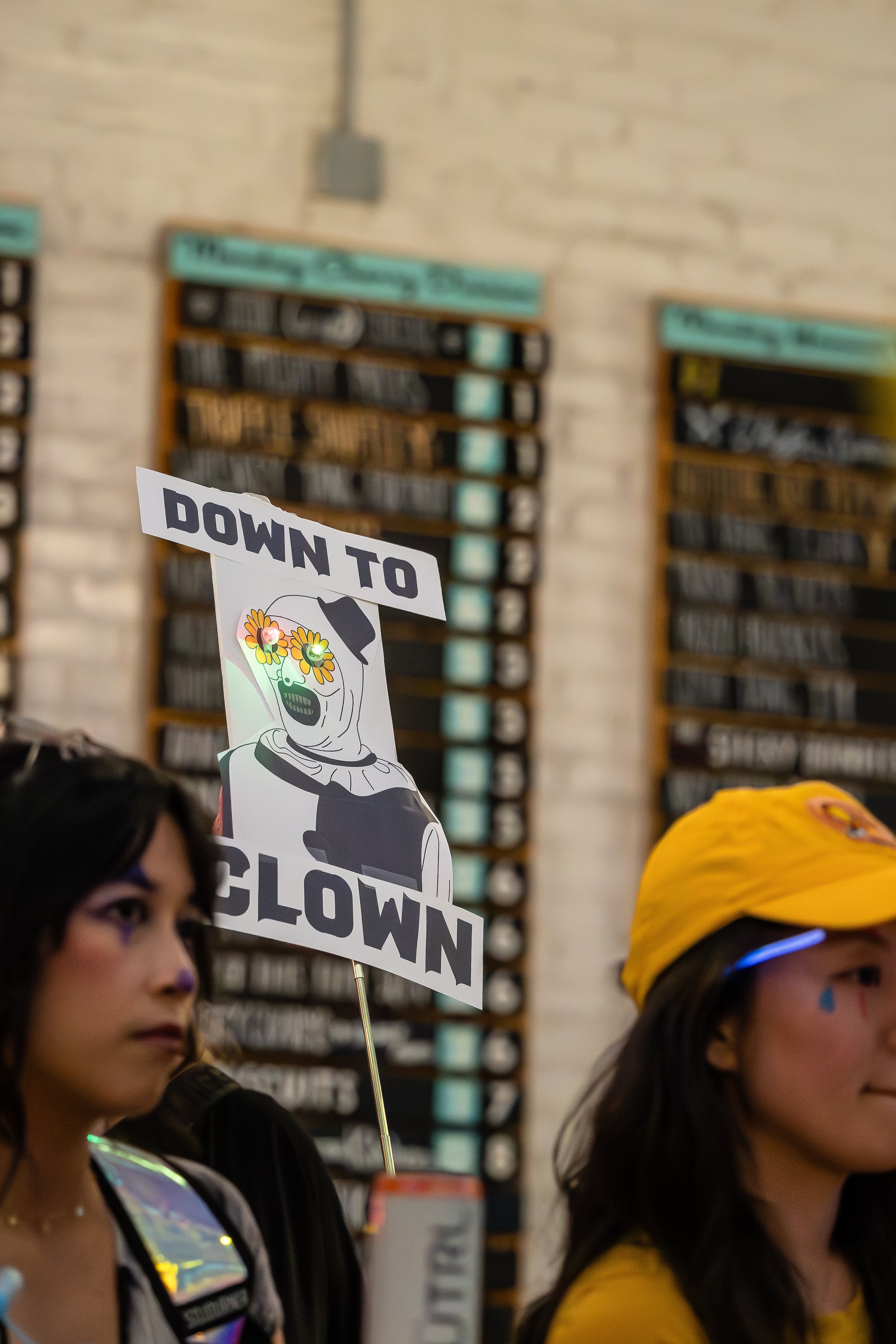 People participating in some event or gathering, with one holding a sign that says "DOWN TO GLOWN" featuring a skeleton with flowers over its eyes. The scene appears to be indoors, with a brick wall and bulletin boards in the background.