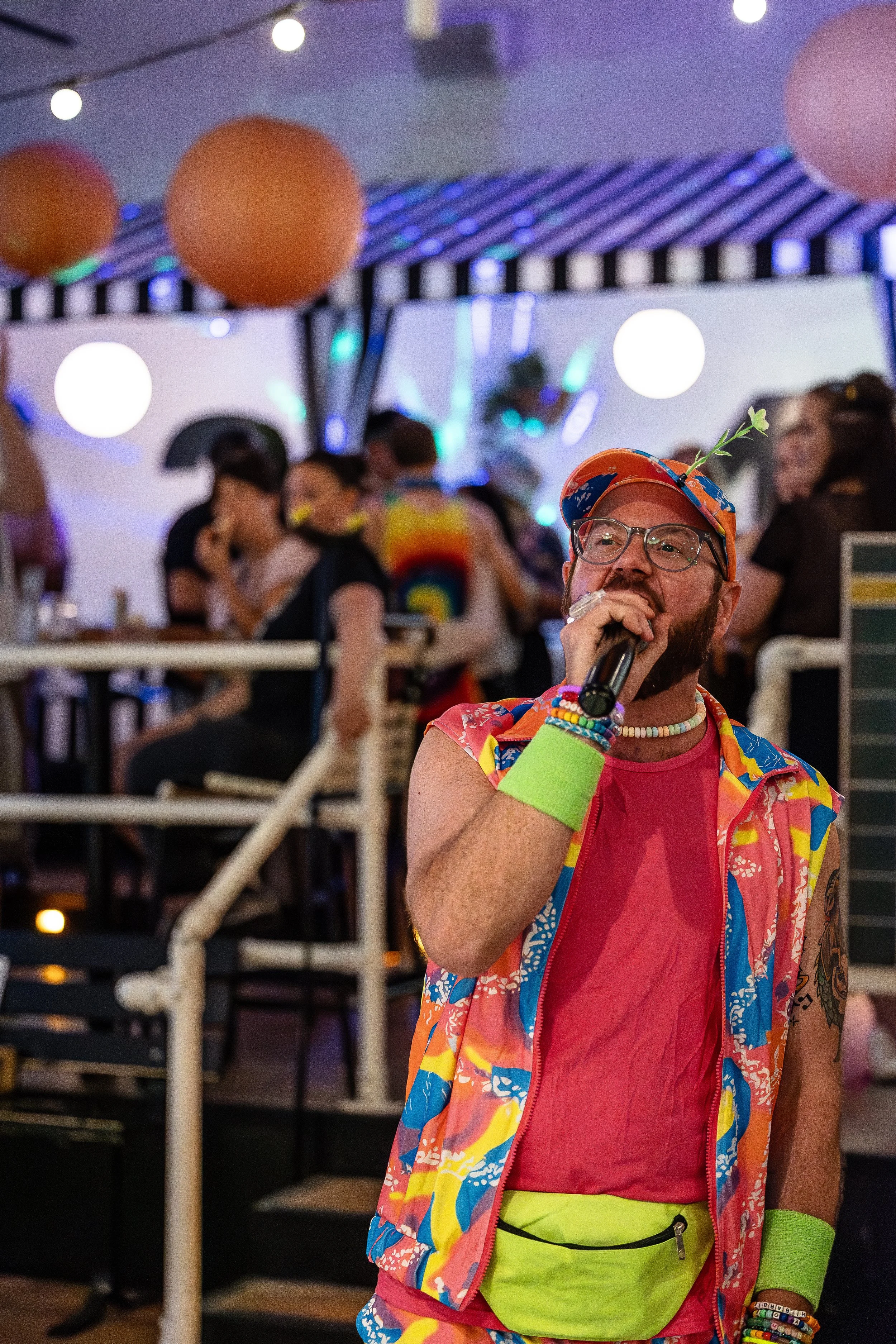 Man dressed in colorful, retro-style clothing singing into a microphone at a lively party with decorations and people dancing in the background.